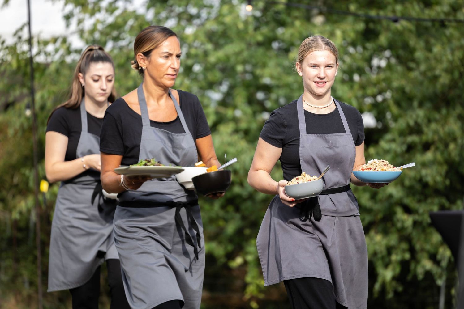 Three servers in gray aprons carry plates while working at an outdoor dining event.