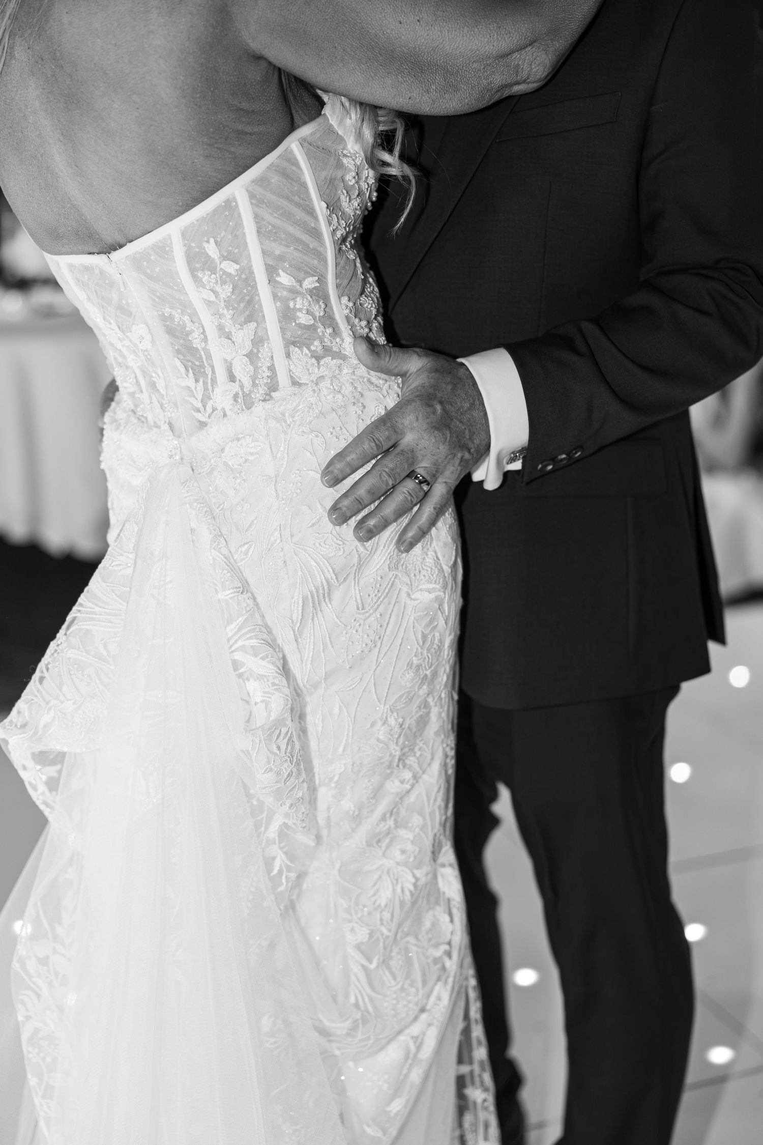 Black and white photo of bride and groom sharing first dance, groom's hand on bride's lace wedding gown waist.