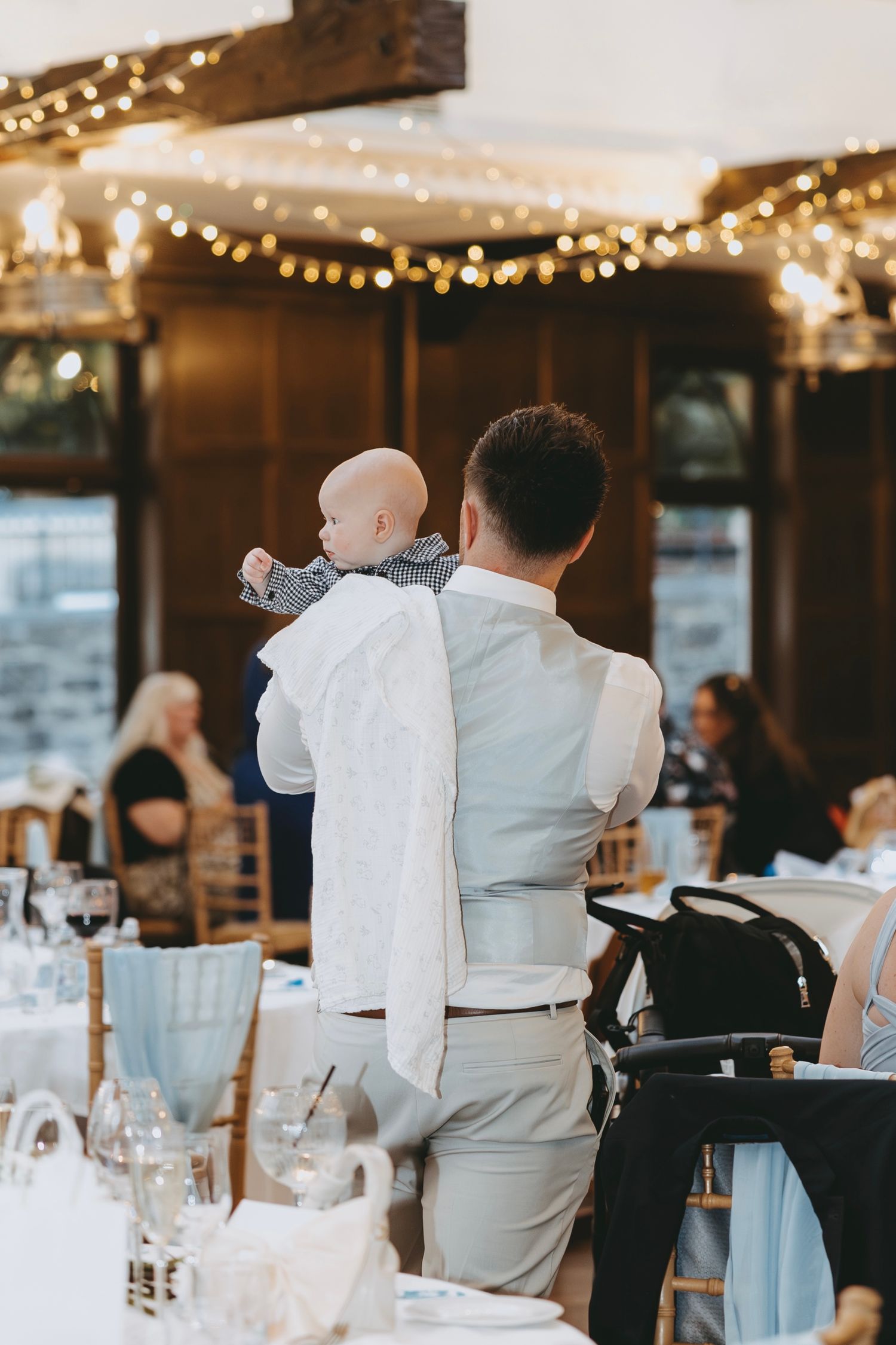 A small child in a white outfit is held up during a wedding reception with string lights in the background.