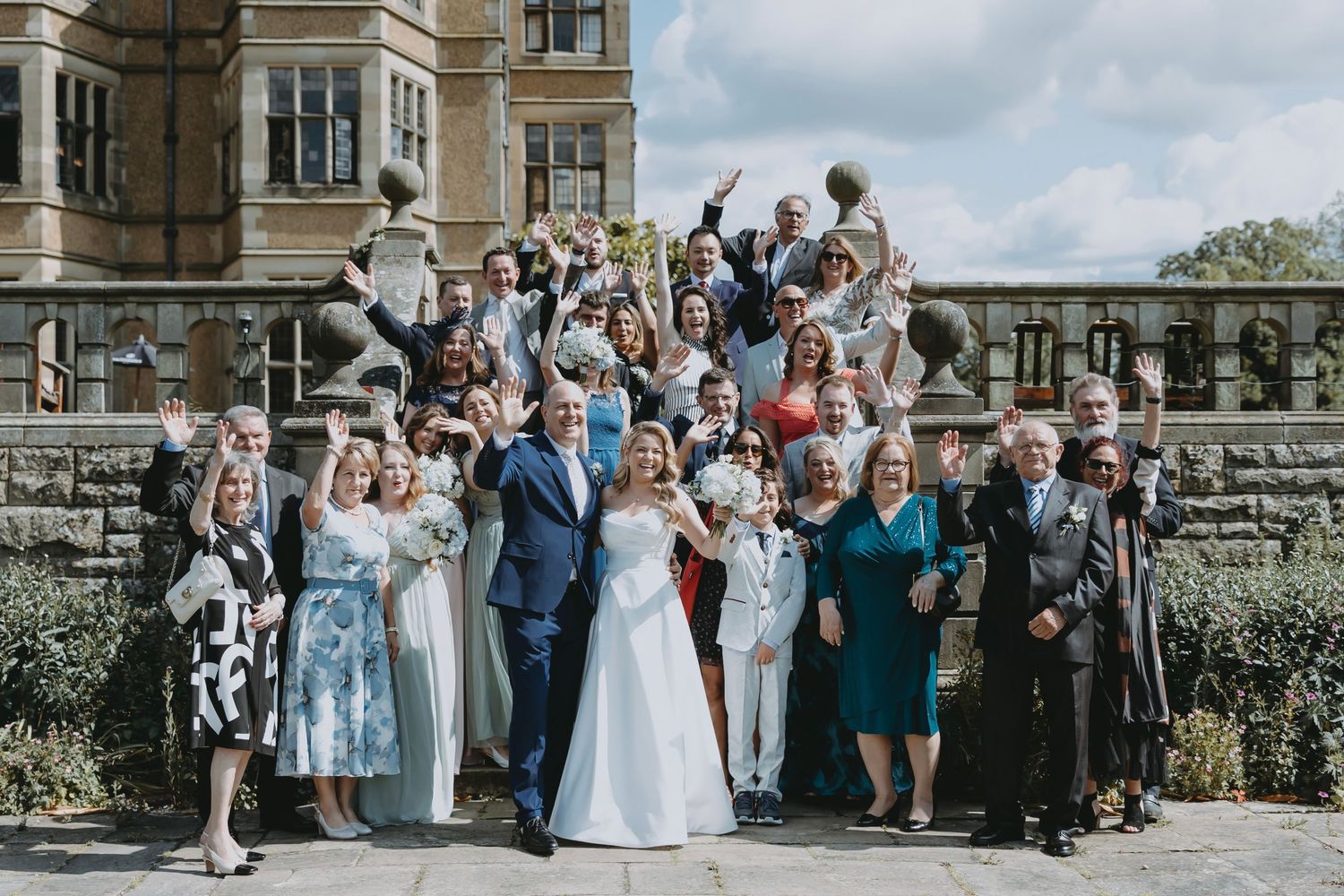 Large group wedding photo with guests celebrating on stone terrace.