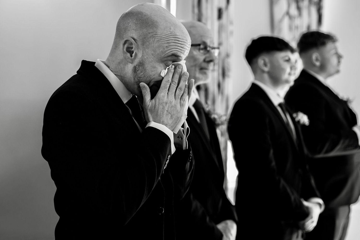 Emotional groom wiping tears during wedding ceremony with groomsmen in background.