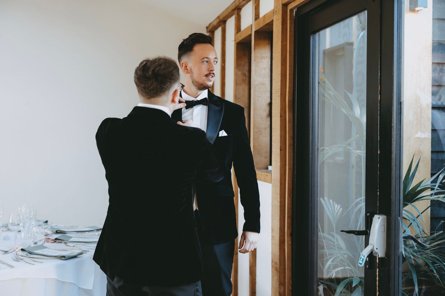 A formal wedding party member adjusts his tie in front of wooden-framed windows.