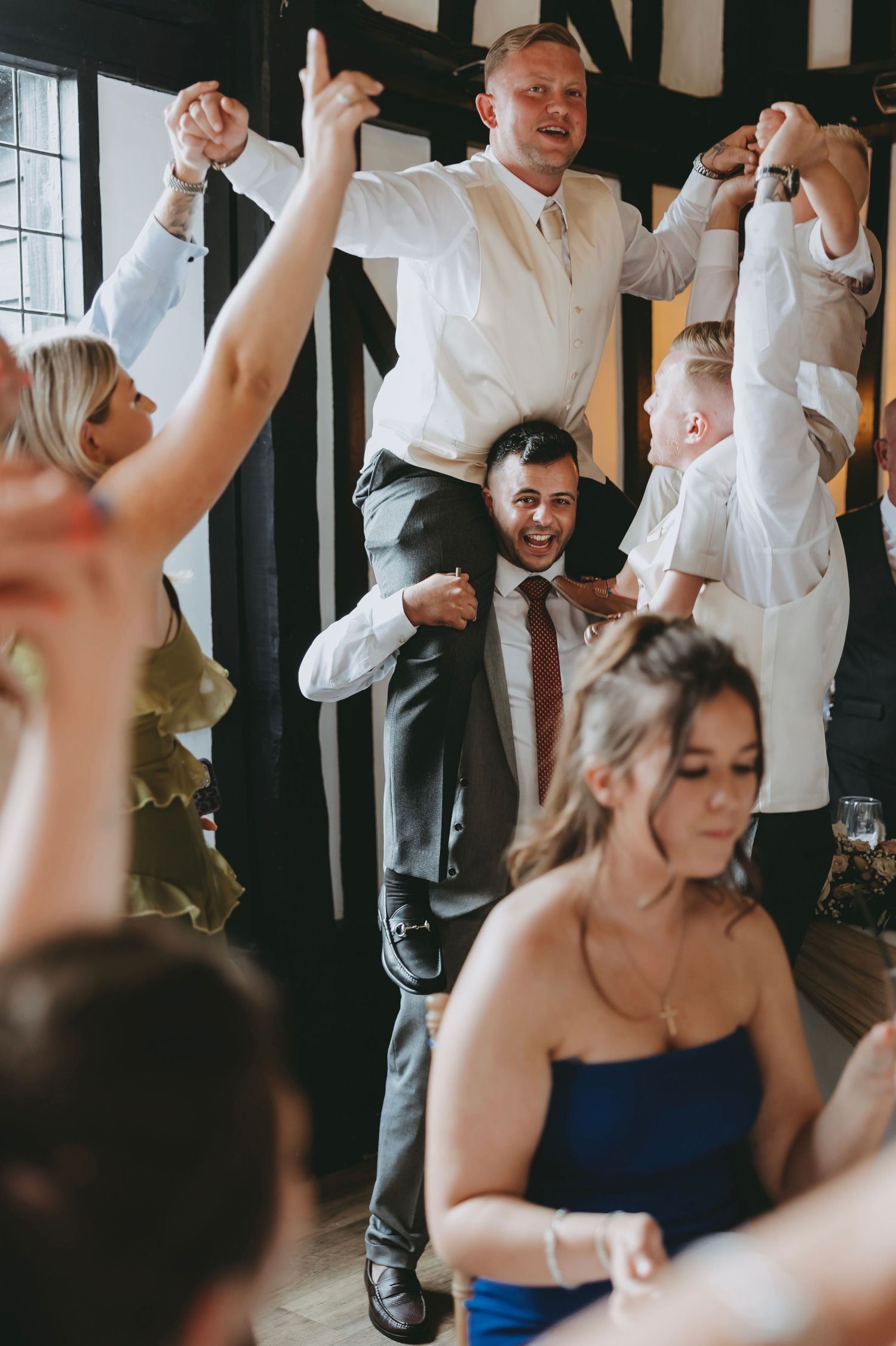 Wedding party dancing with enthusiasm at an indoor reception with large windows.