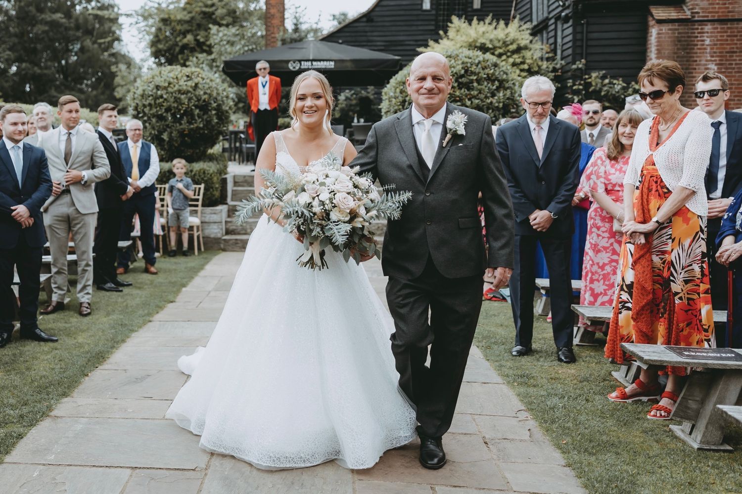 Outdoor wedding ceremony with stone walkway and flowering bushes in background.
