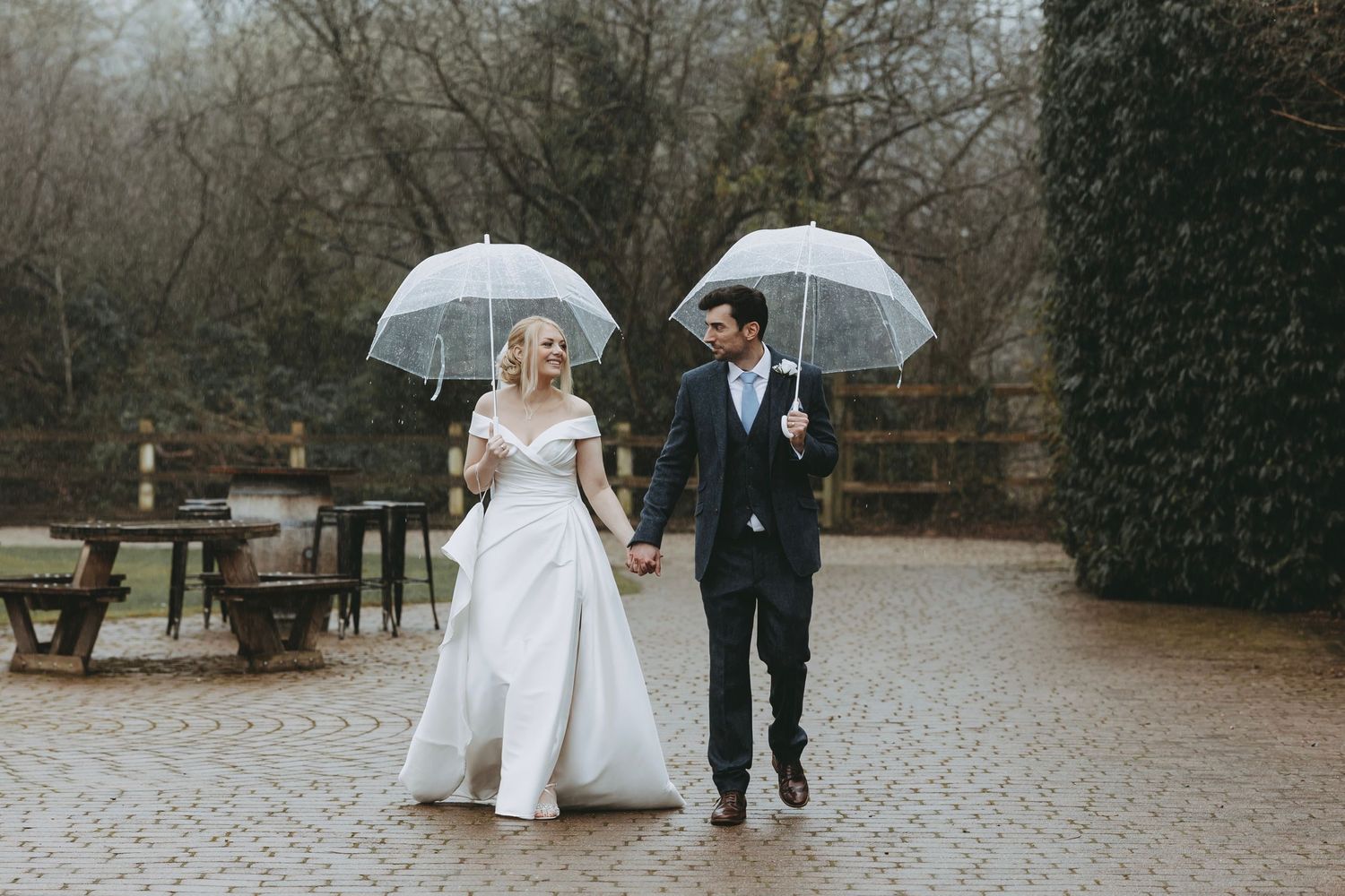 Couple walking together under clear umbrellas on a rainy cobblestone path.