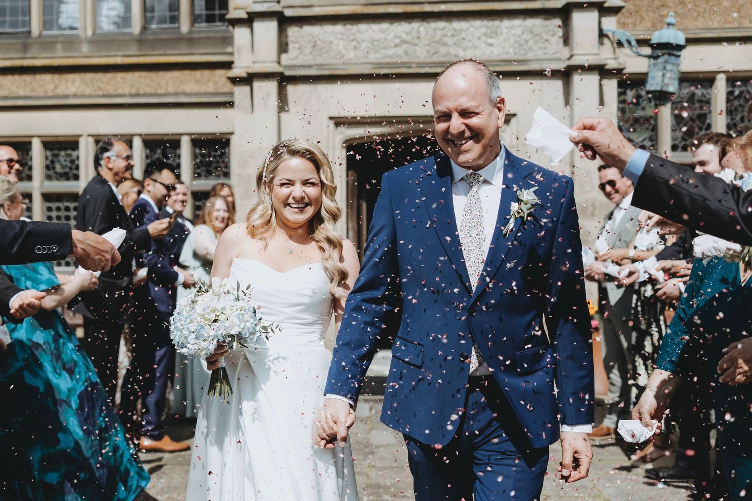 Wedding celebration with guests throwing confetti outside a historic stone building.