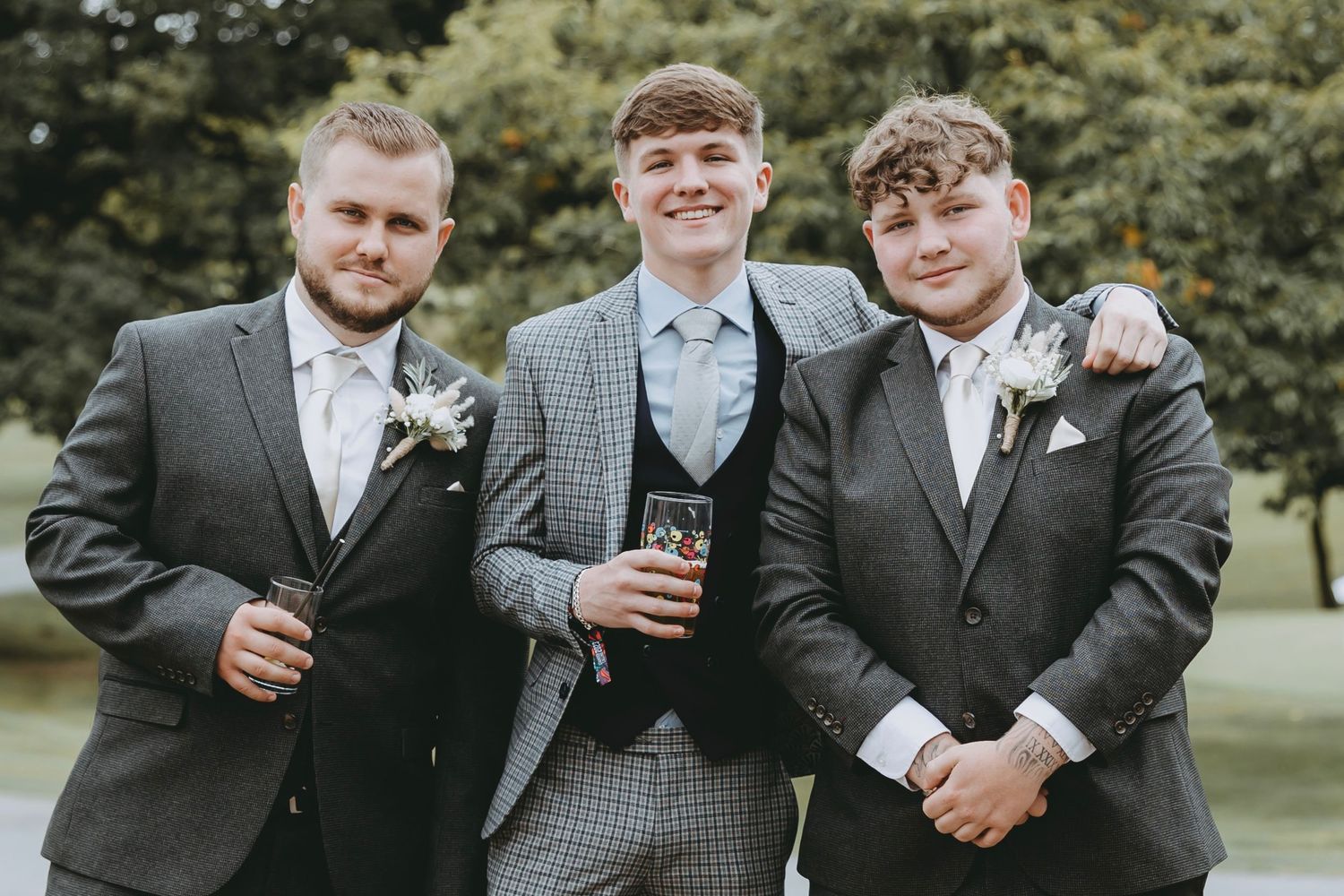 Groomsmen in gray suits share drinks and laughs at an outdoor wedding.