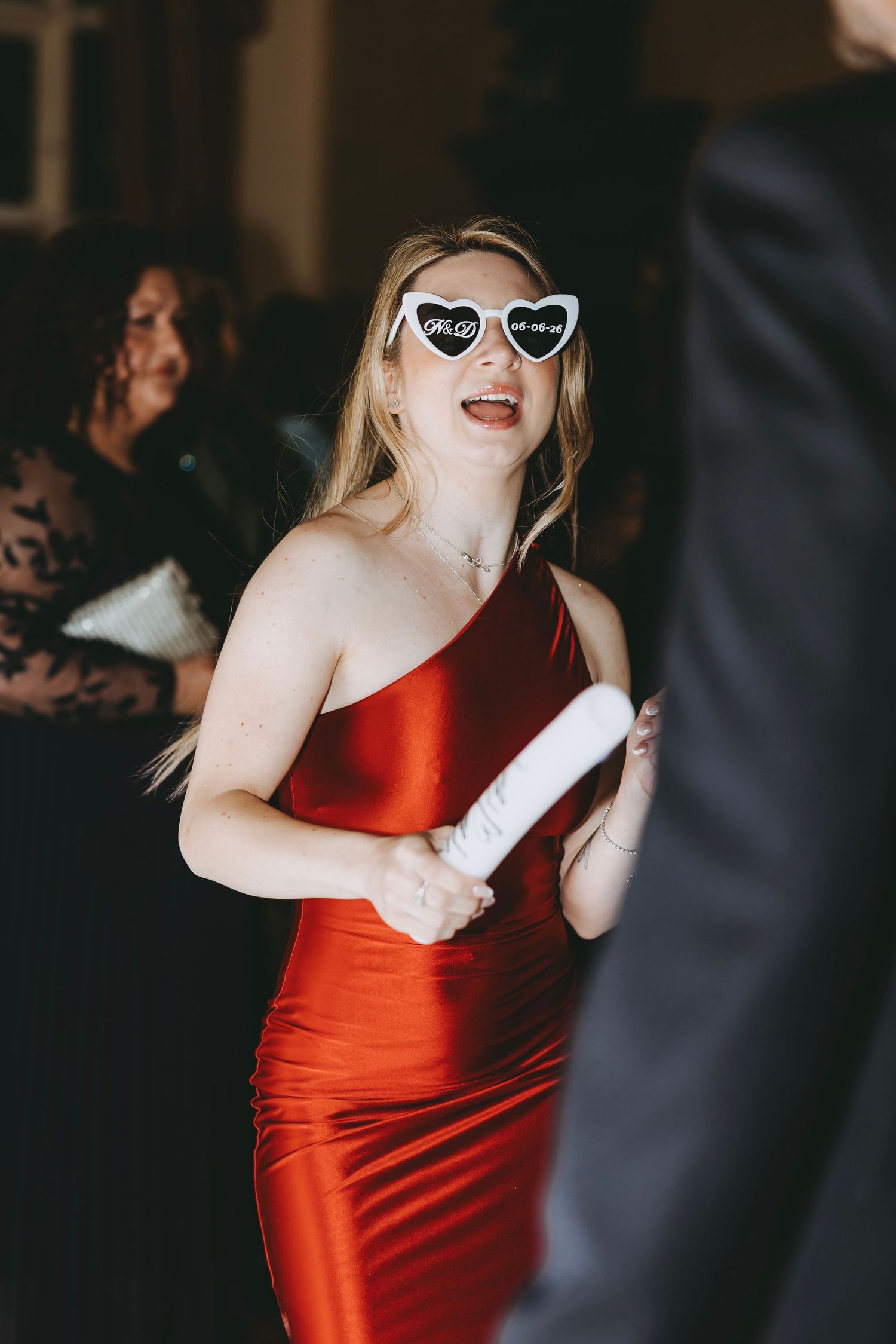 Blonde woman in red satin dress wearing heart-shaped novelty sunglasses dancing at a wedding reception.