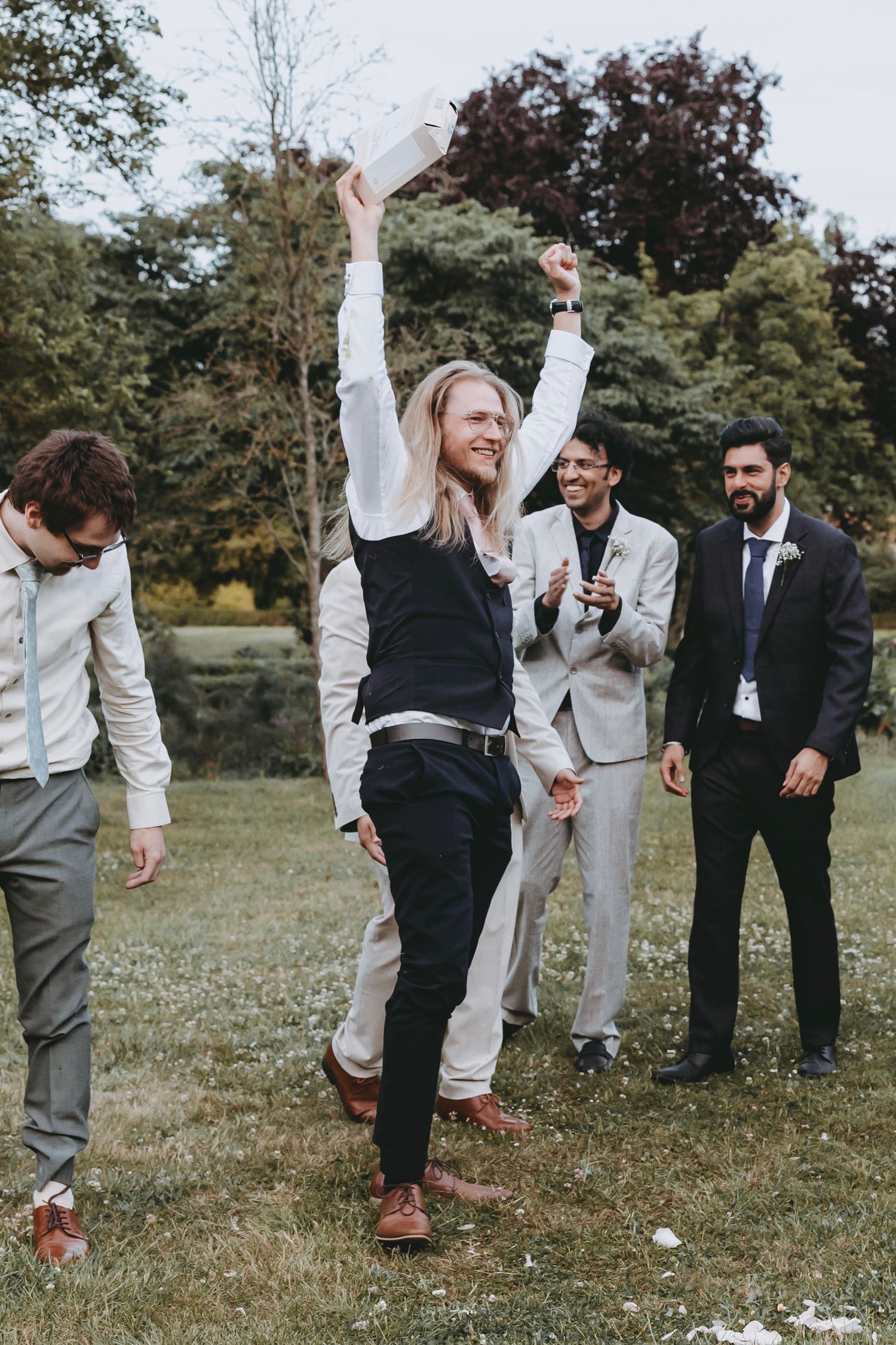 Long-haired man celebrates outdoors at wedding, raising arms in victory while groomsmen cheer on green lawn.