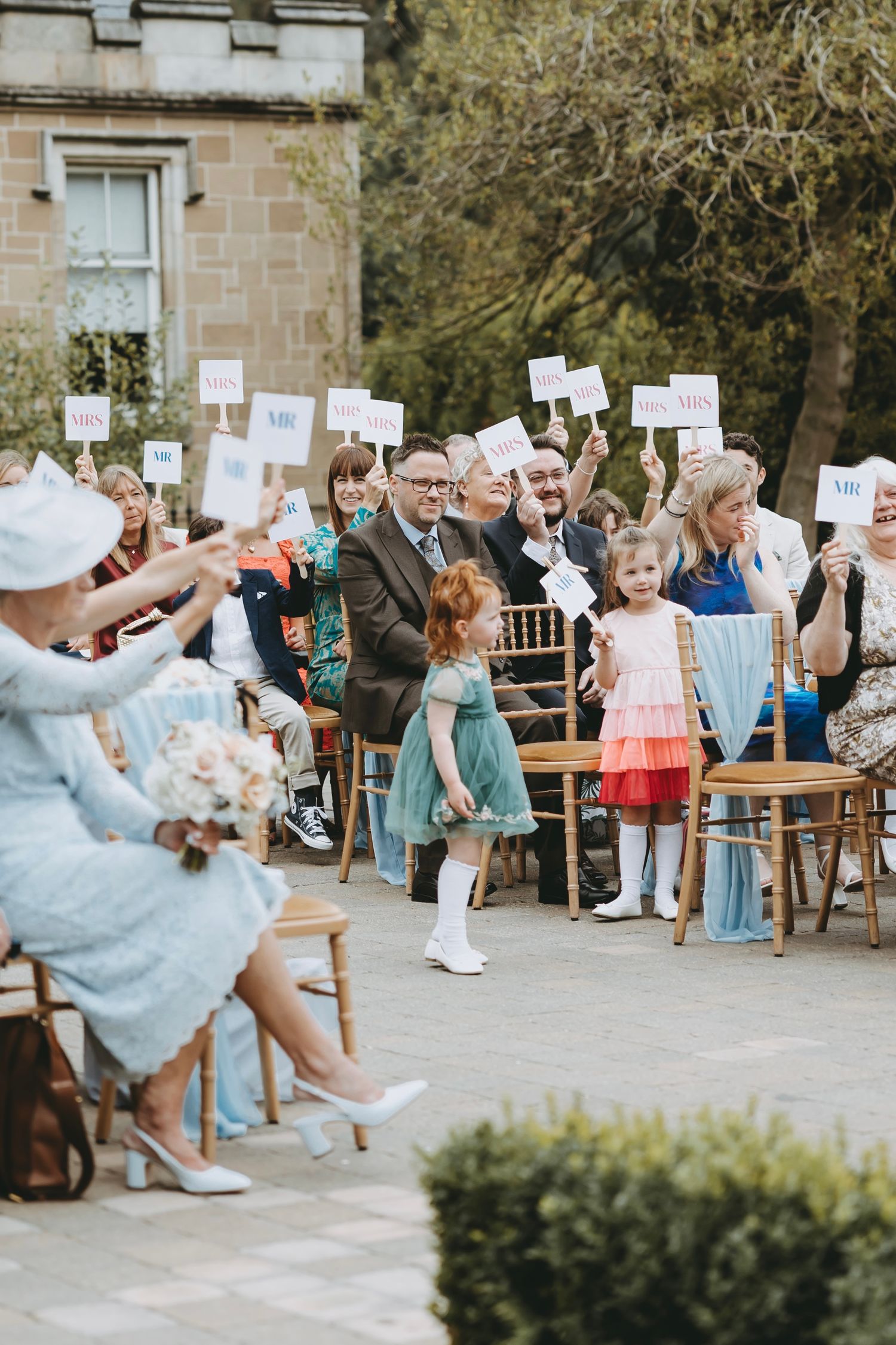 Wedding party members hold up signs while seated in wooden chairs during an outdoor game.