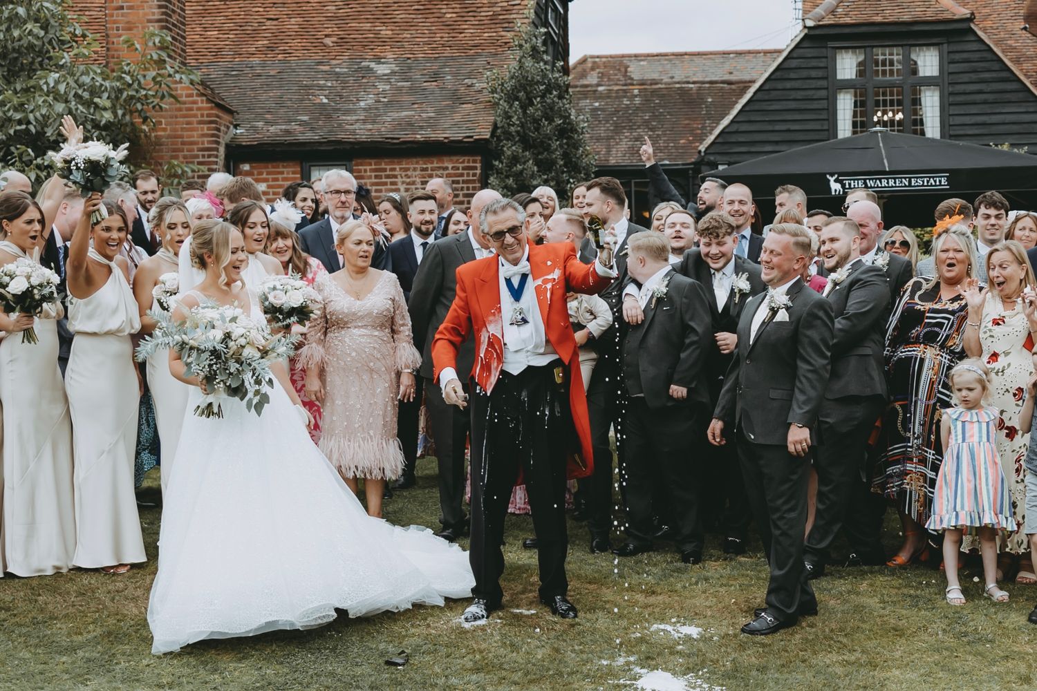 Large wedding party gathering outdoors with guests in formal attire against a brick building backdrop.