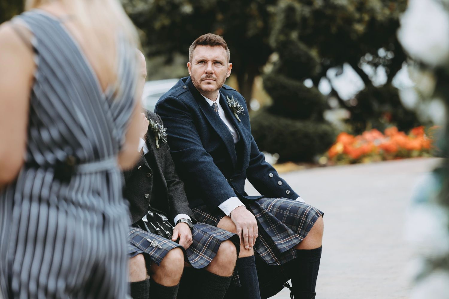 Wedding guests in striped dress and traditional Scottish attire at outdoor ceremony.