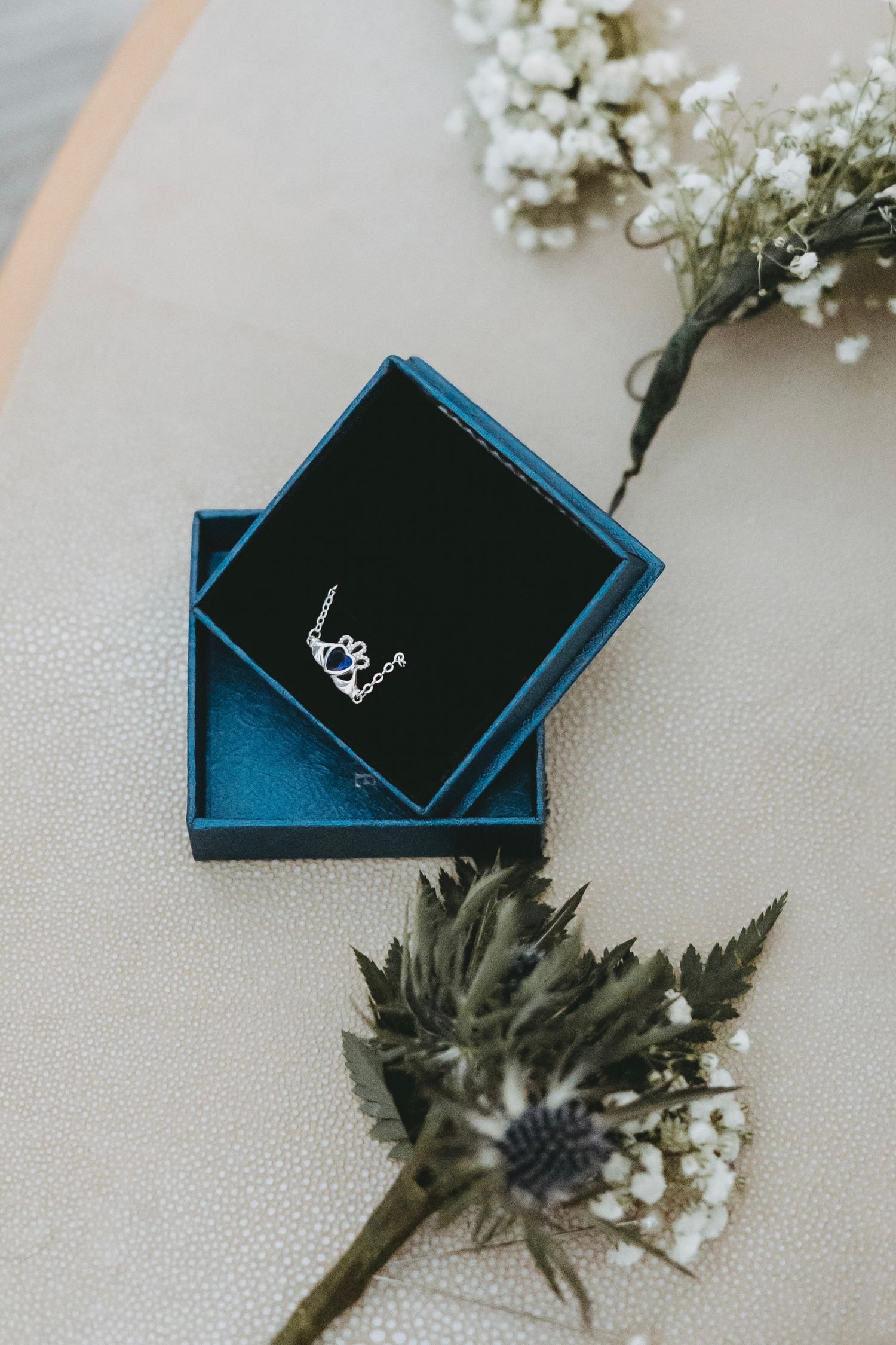 Small blue velvet jewelry box with white flowers and greenery displayed on light surface.