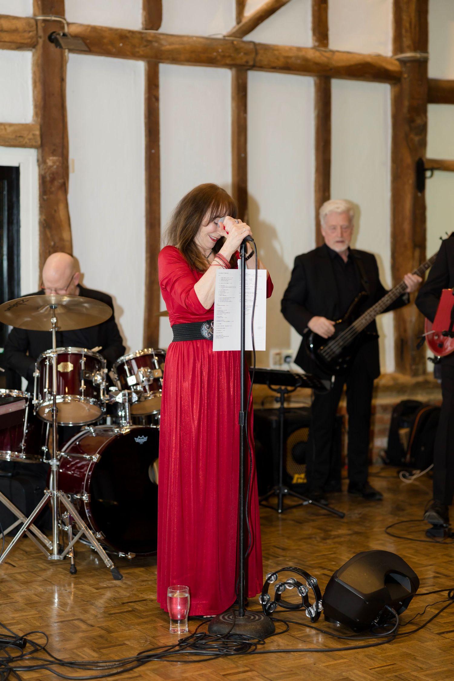 A performer in a red dress sings on stage with a band setup including drums and instruments in a rustic venue.