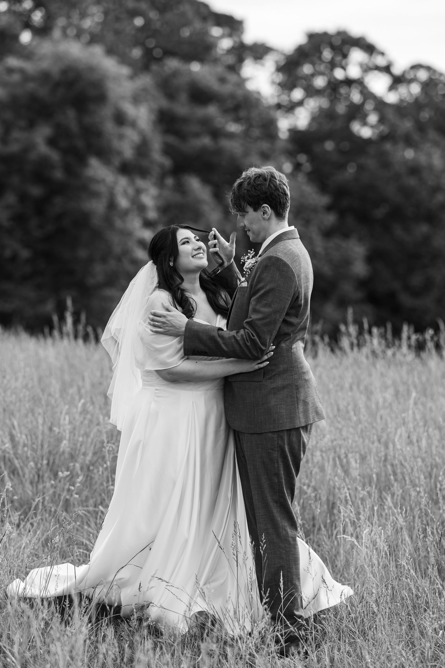 Black and white photo of a bride and groom embracing in a meadow, bride smiling in white gown with veil.