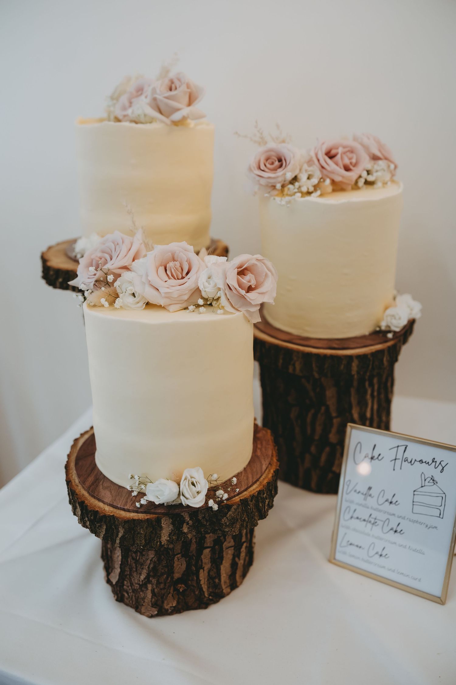 White wedding cake decorated with pink roses displayed on rustic wood slice stands.
