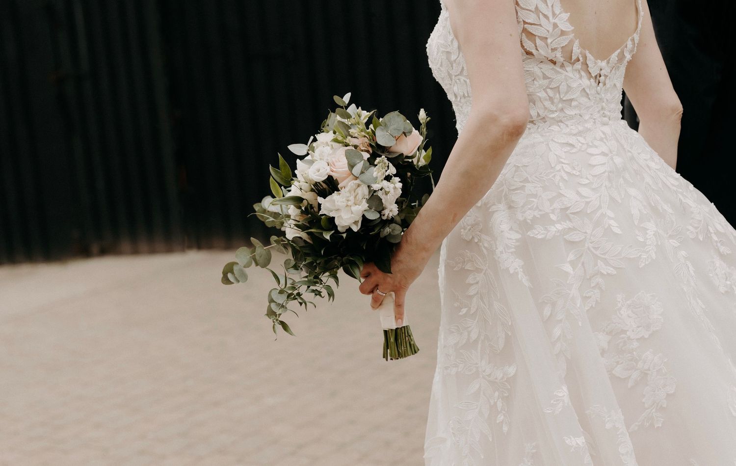 A wedding dress detail showing intricate lace and beading patterns against a dark background.