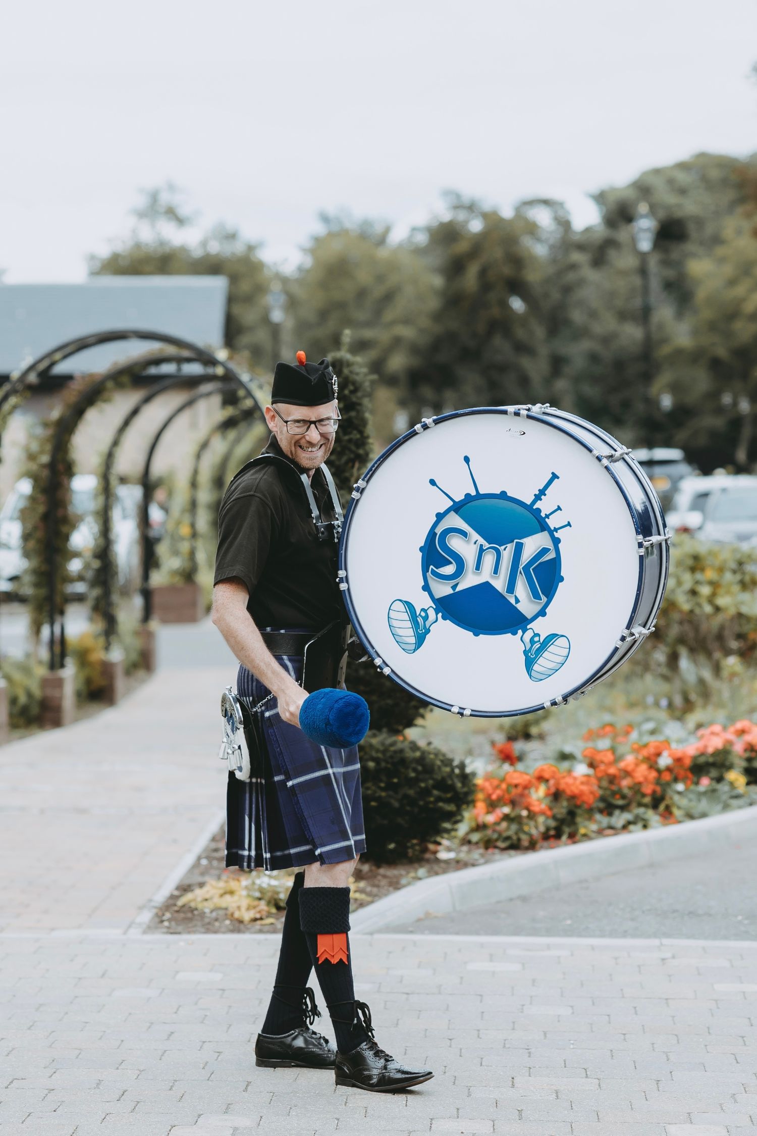 Bagpiper carrying decorated drum case across stone courtyard.