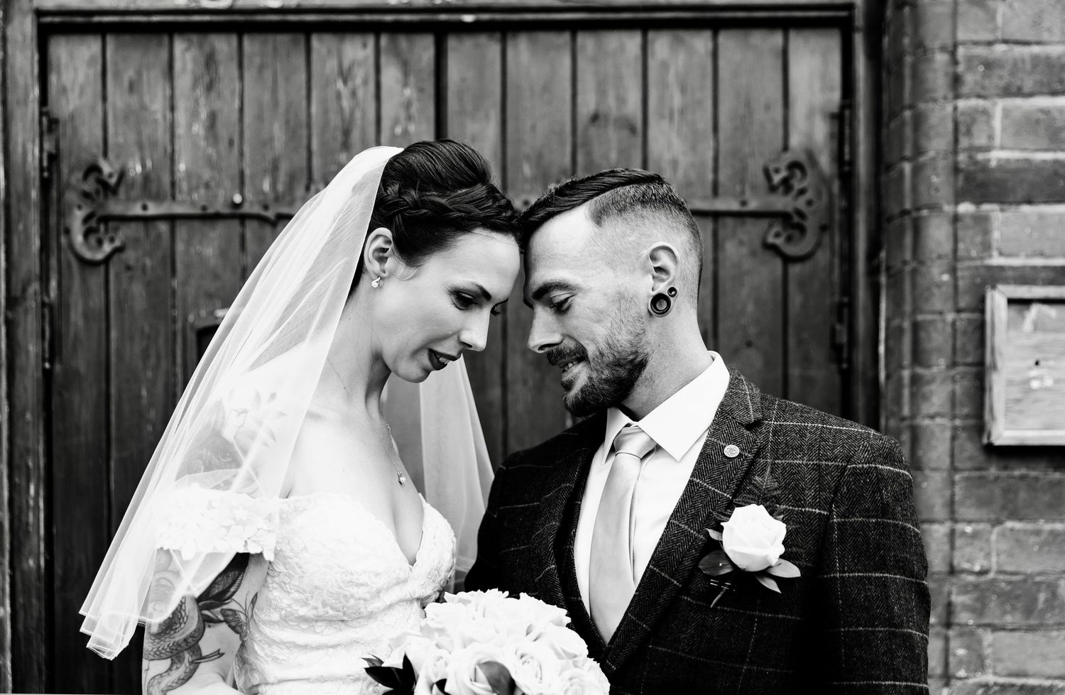 Black and white wedding portrait showing tender romantic moment against brick wall backdrop.