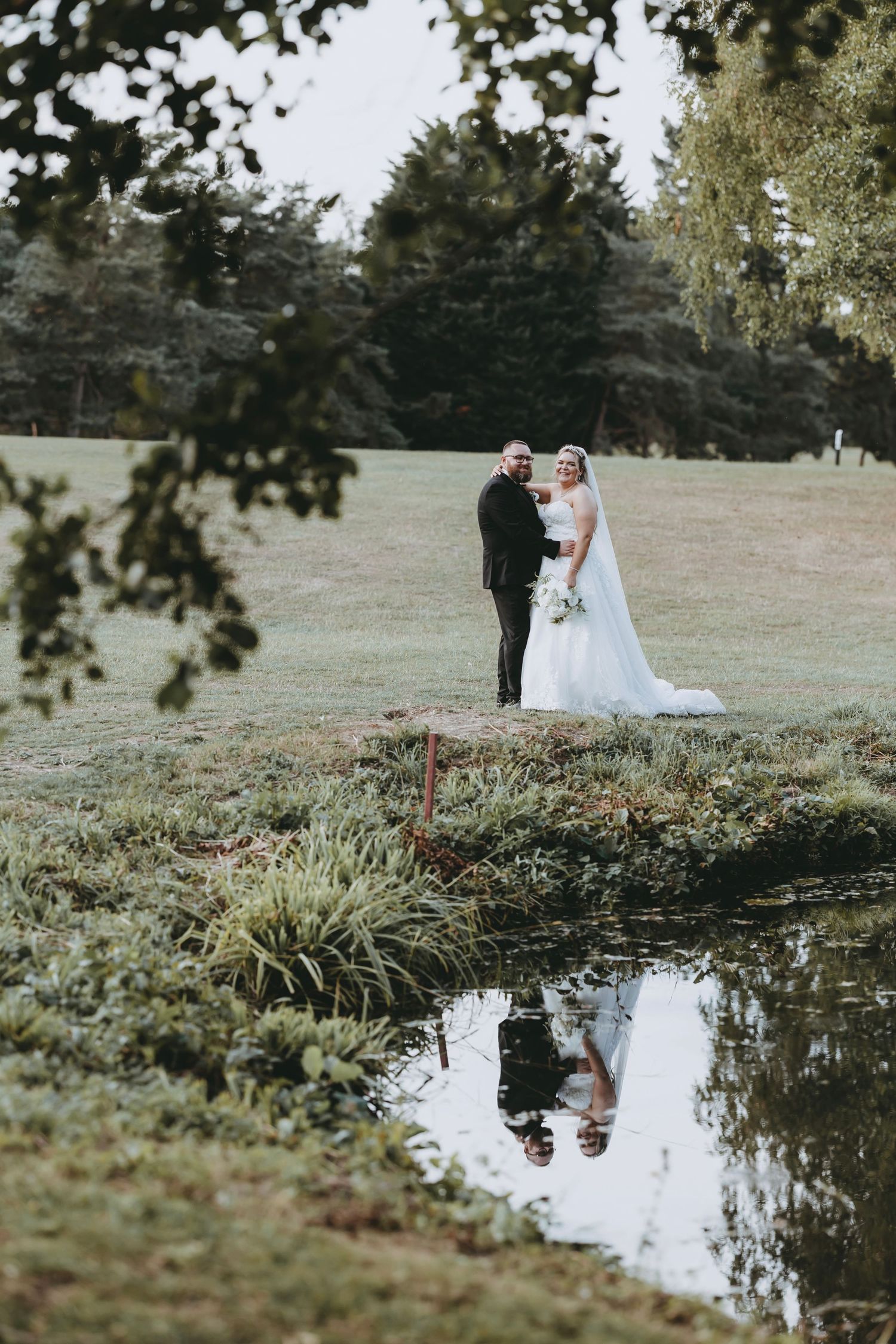Wedding couple reflected in a serene pond surrounded by natural landscaping.