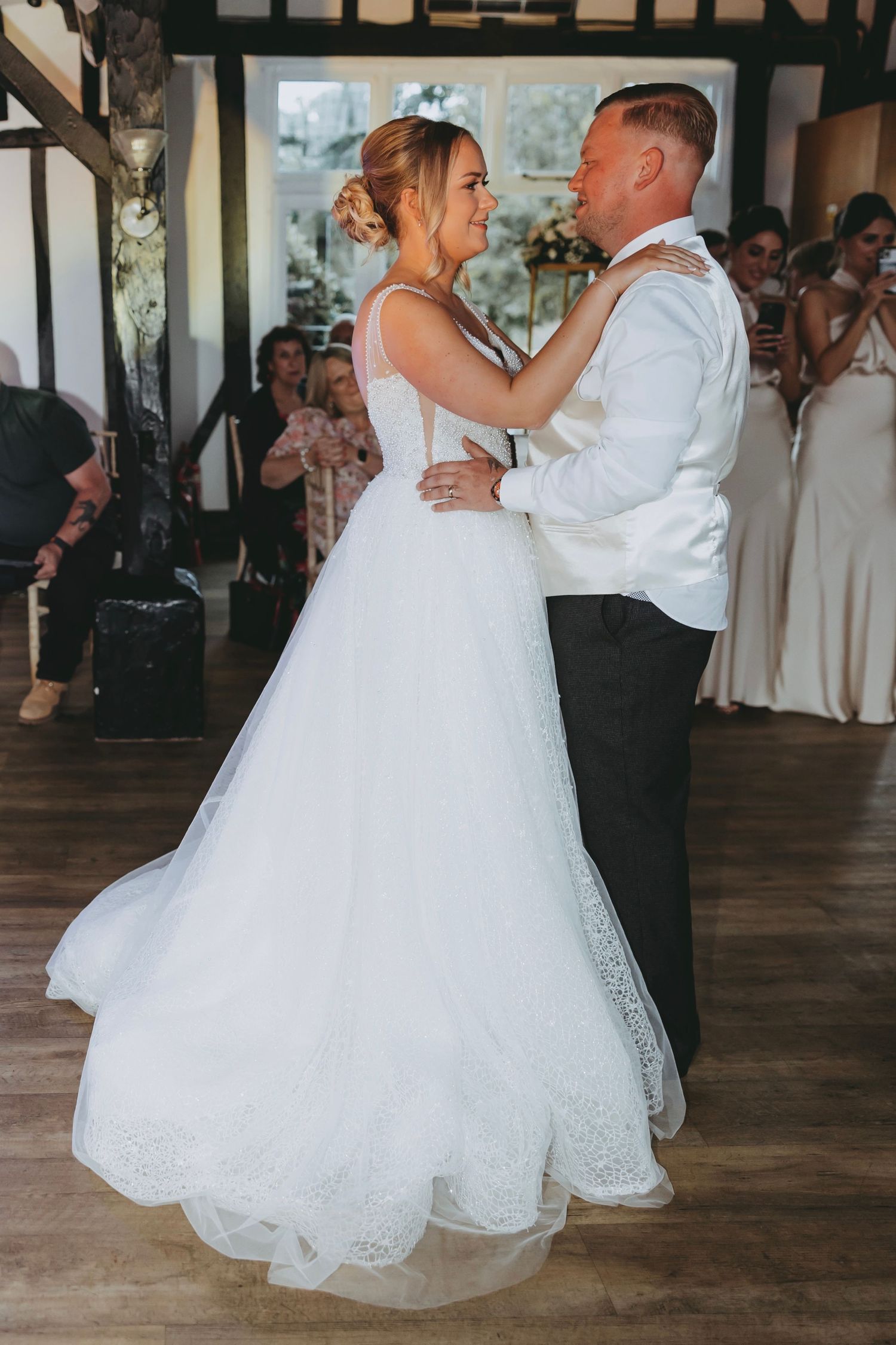 Newlyweds dancing together in a ballroom with romantic lighting and guests in the background.