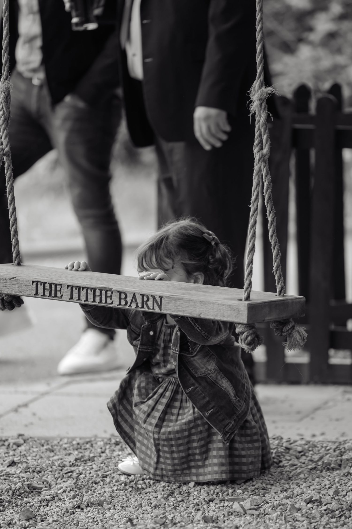 Black and white photo of a young girl peeking over a wooden swing labeled The Tithe Barn at an outdoor venue.