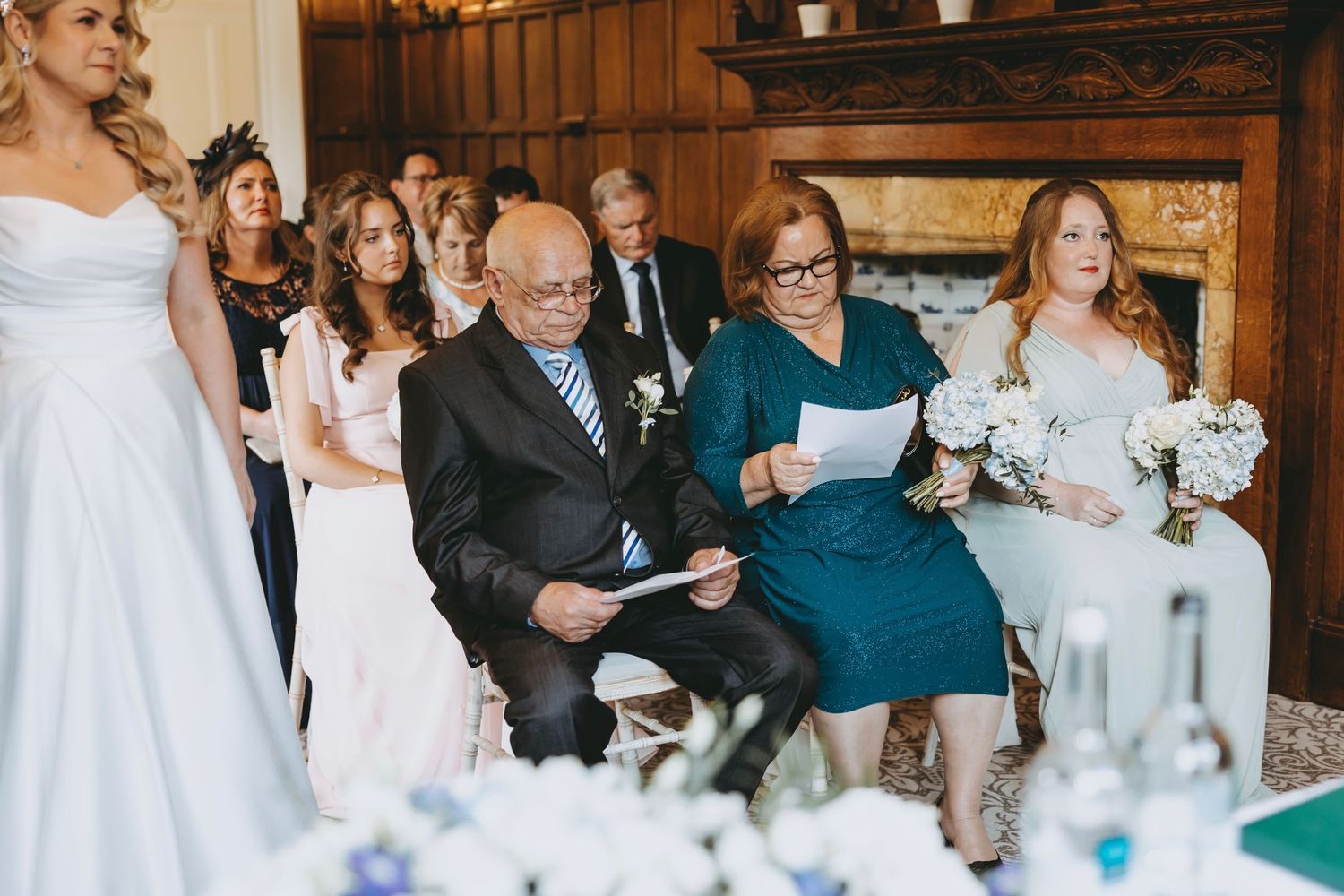 Wedding guests seated in formal attire watching ceremony in traditional wood-paneled venue.