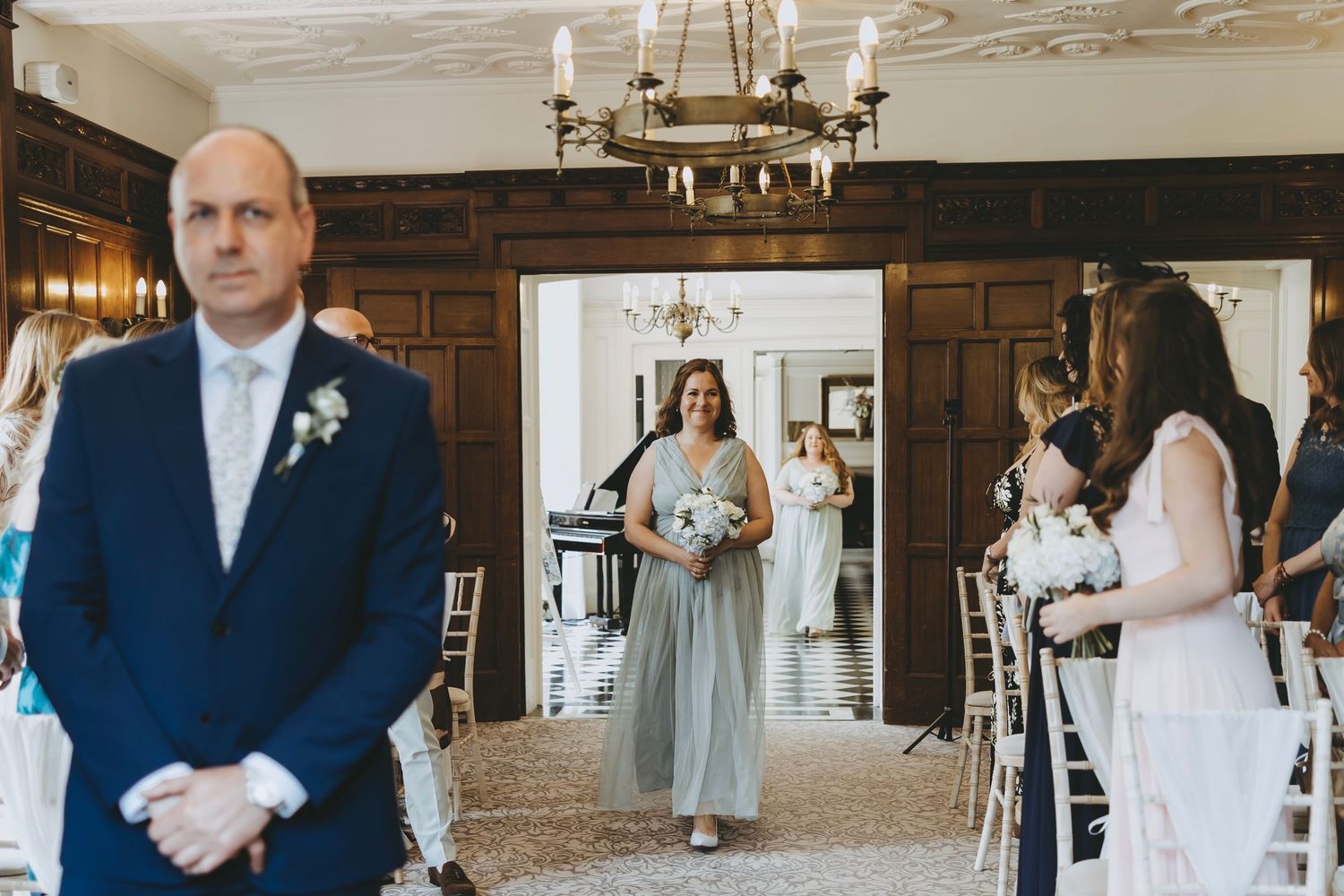 Wedding ceremony in an elegant wood-paneled room with chandelier and attendants in light blue and white dresses.