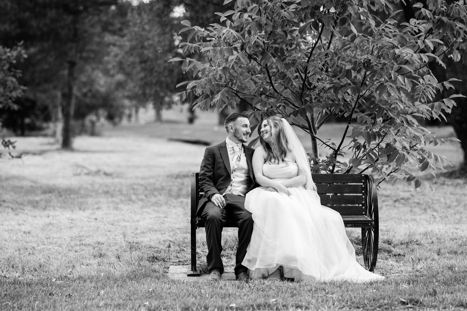 A couple sits together on a park bench in black and white wedding attire during an outdoor photoshoot.