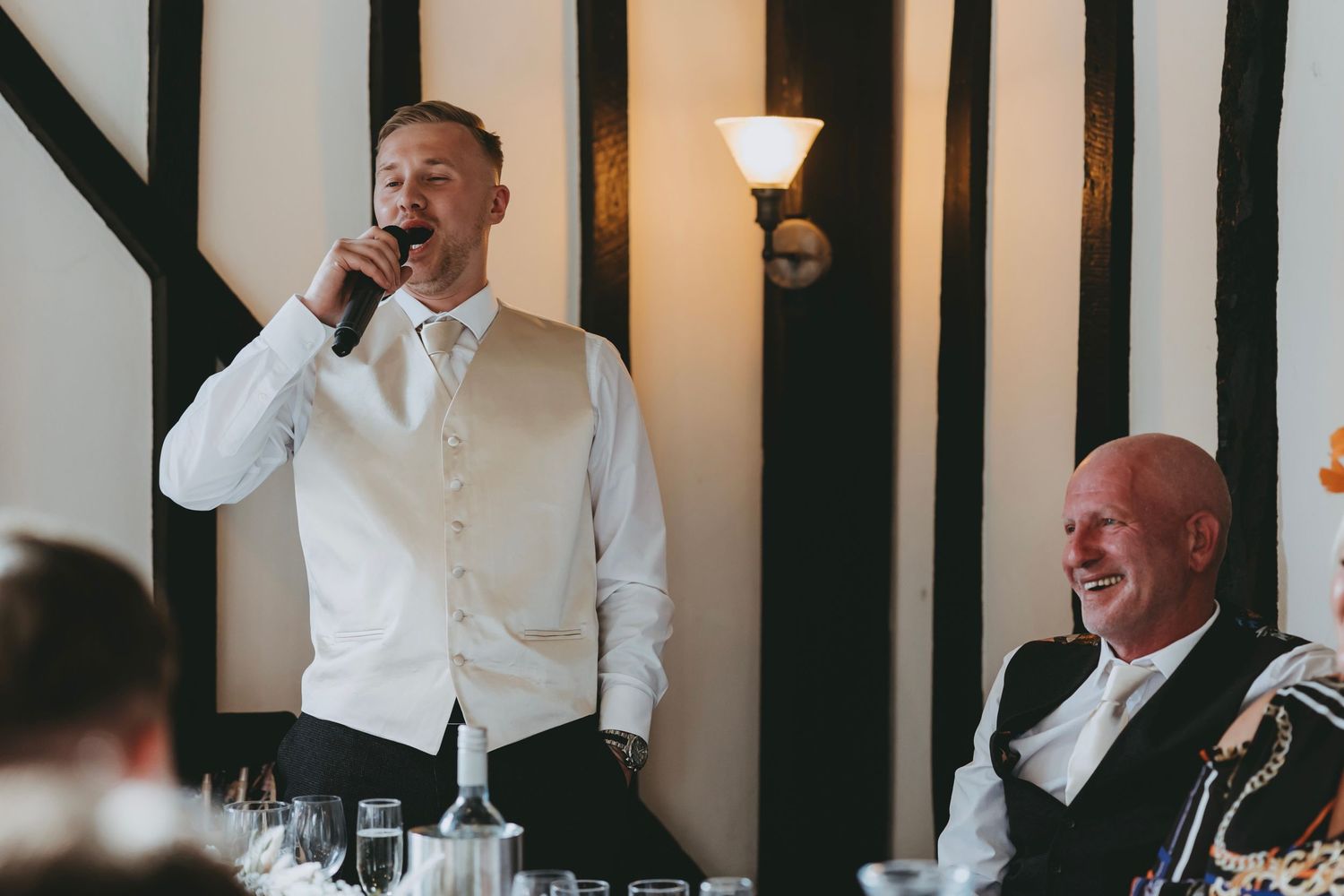 Wedding guests laugh and react emotionally during speeches at a reception with black and white timber beam decor.