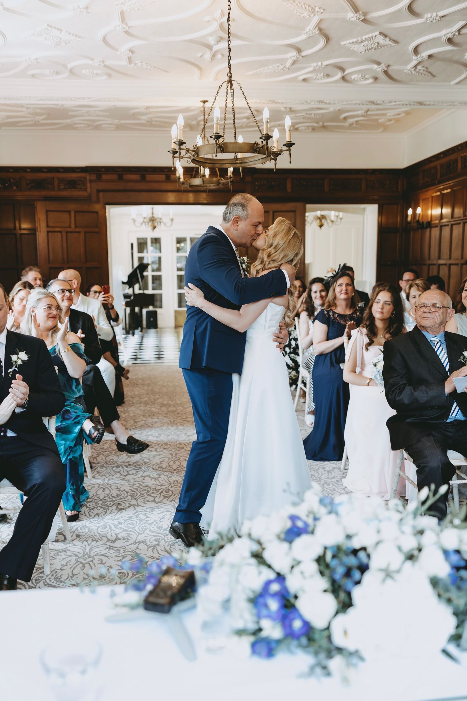 Wedding guests during the ceremony at Fanhams Hall, Ware, Herts with ornate white ceiling details and blue floral decorations.