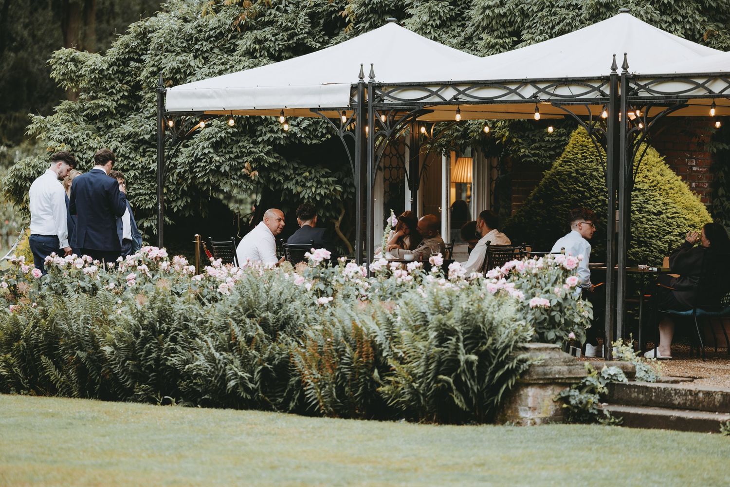 Elegant outdoor garden party with white gazebos, fairy lights, and guests mingling among lush floral arrangements.