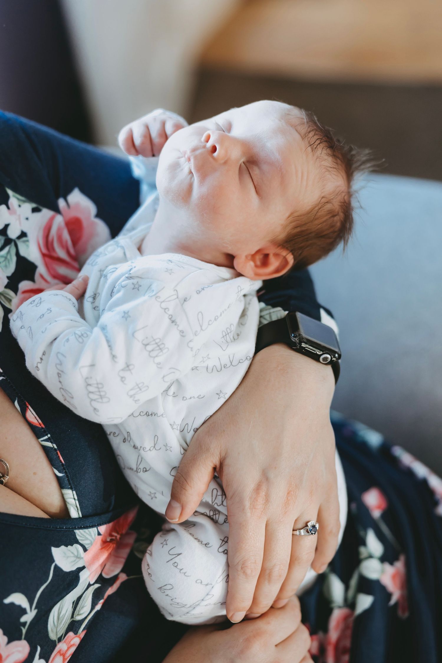 A sleeping newborn baby in a white patterned onesie rests peacefully on a floral dress while being cradled.