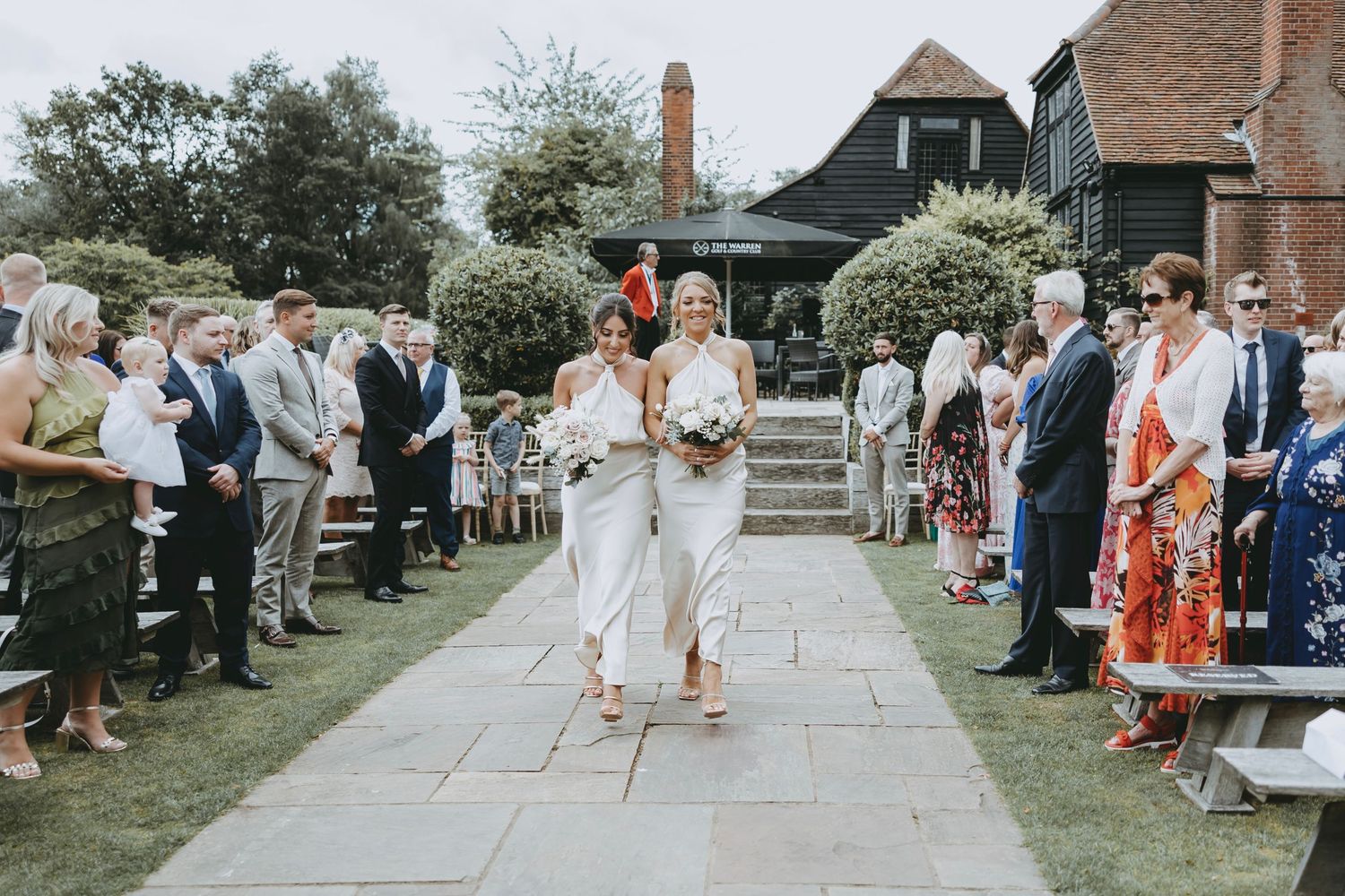 Wedding guests gather on a garden path lined with trees and bushes during an outdoor celebration.