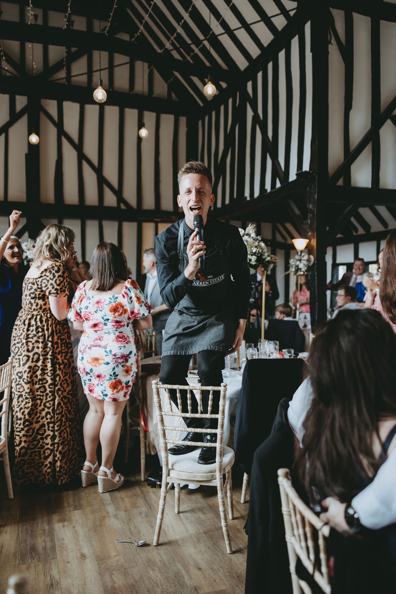 Wedding guests dance and celebrate in a rustic venue with exposed wooden beams and white chairs.