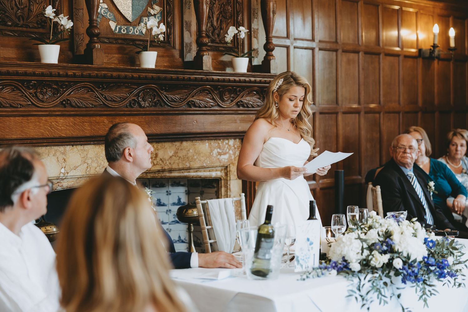 Wedding reception speech moment with elegant table settings and floral decorations in wood-paneled room.