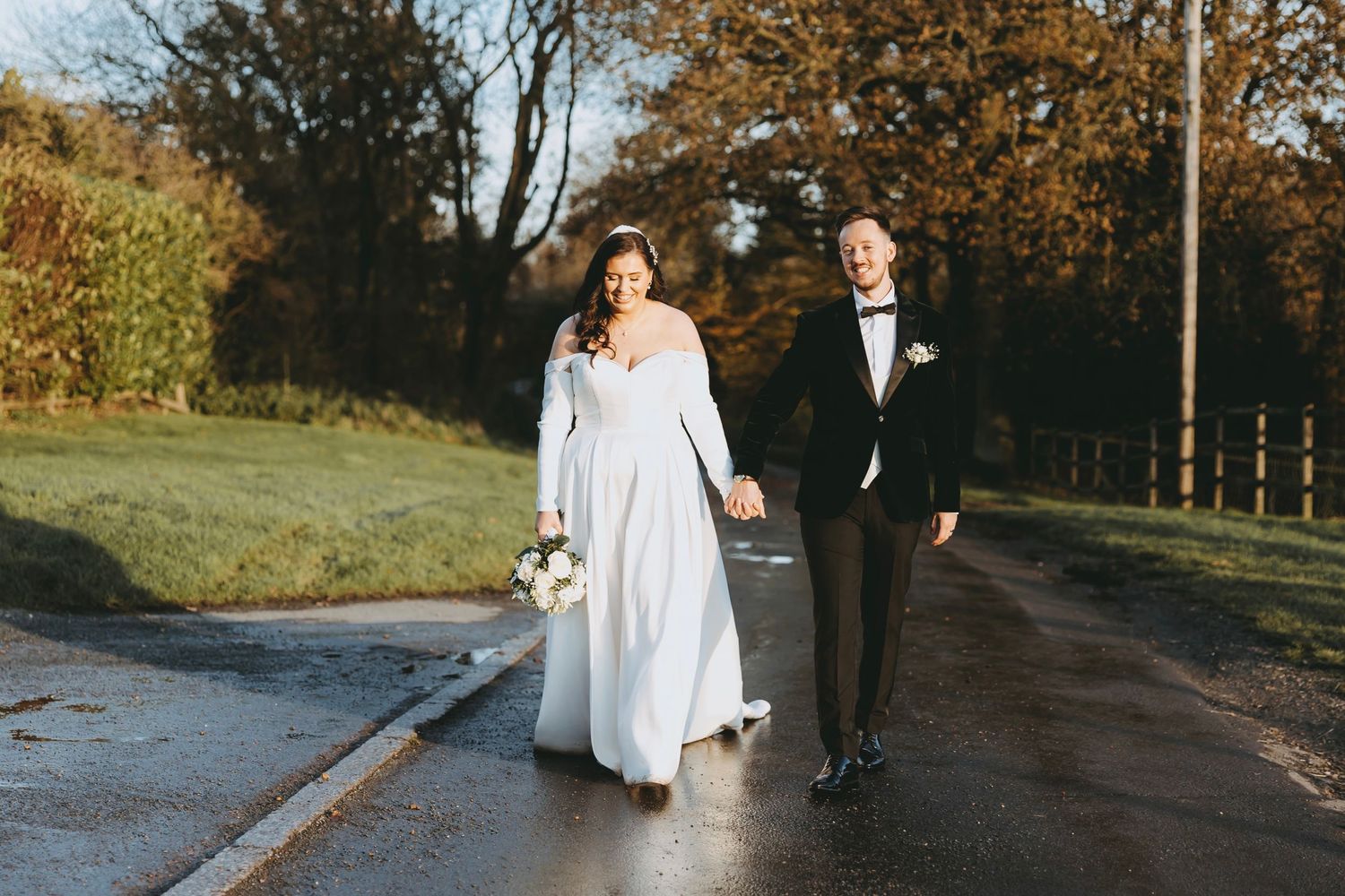 Newlywed couple walks along a tree-lined autumn path during their outdoor wedding photoshoot.