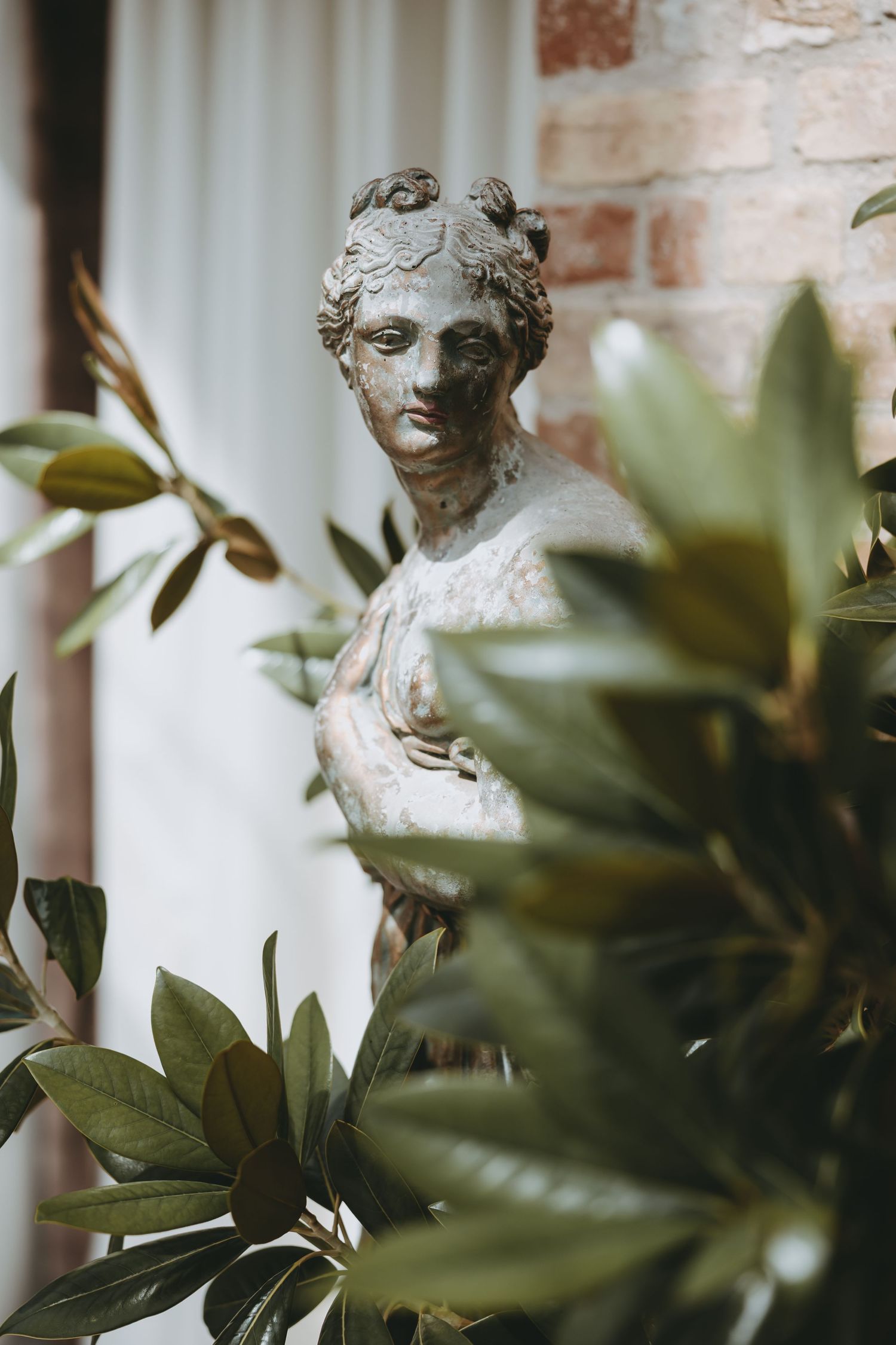 Weathered classical stone bust statue surrounded by lush green leaves against a rustic brick wall backdrop.