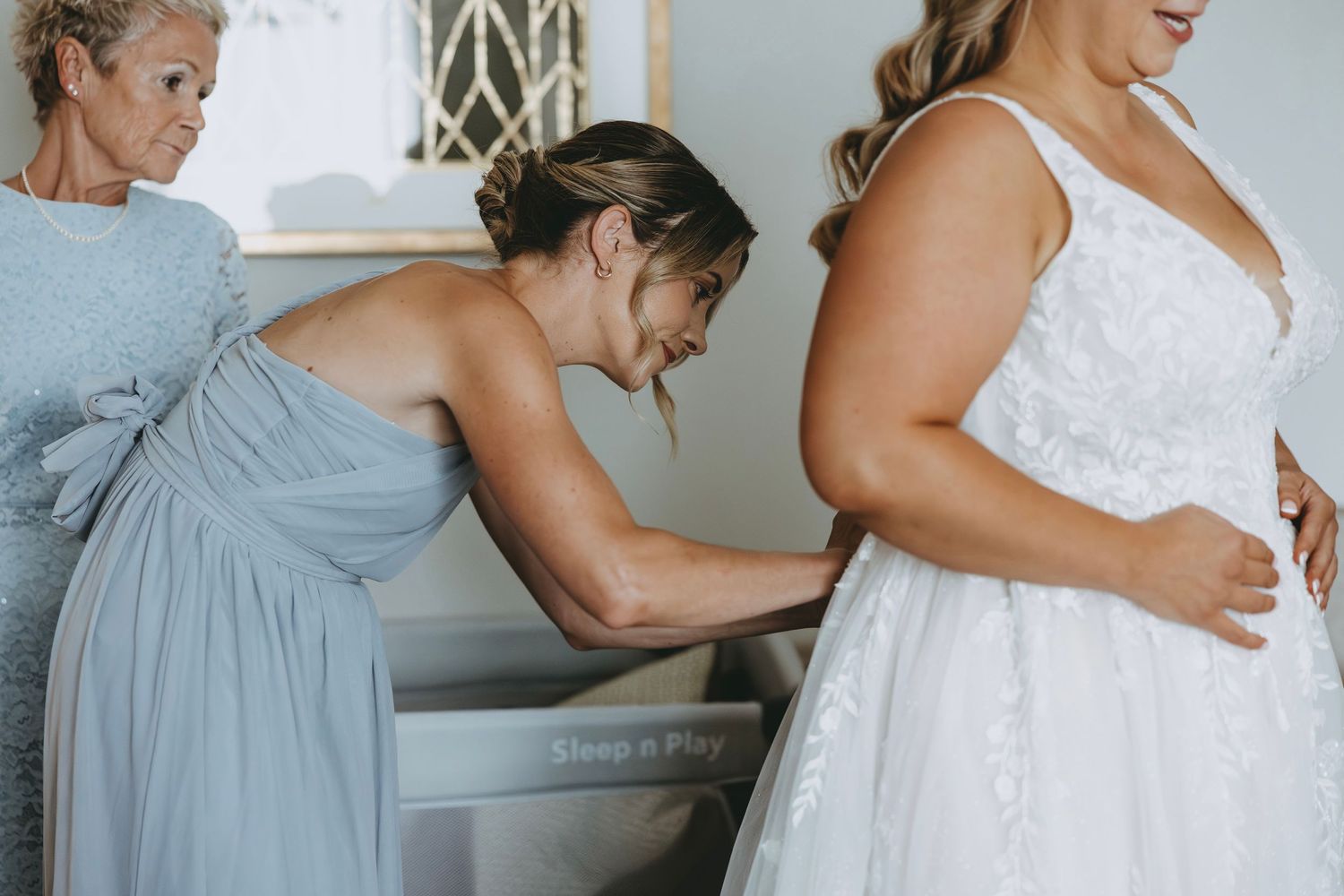 Wedding party members assist with final dress adjustments in a light-filled room.