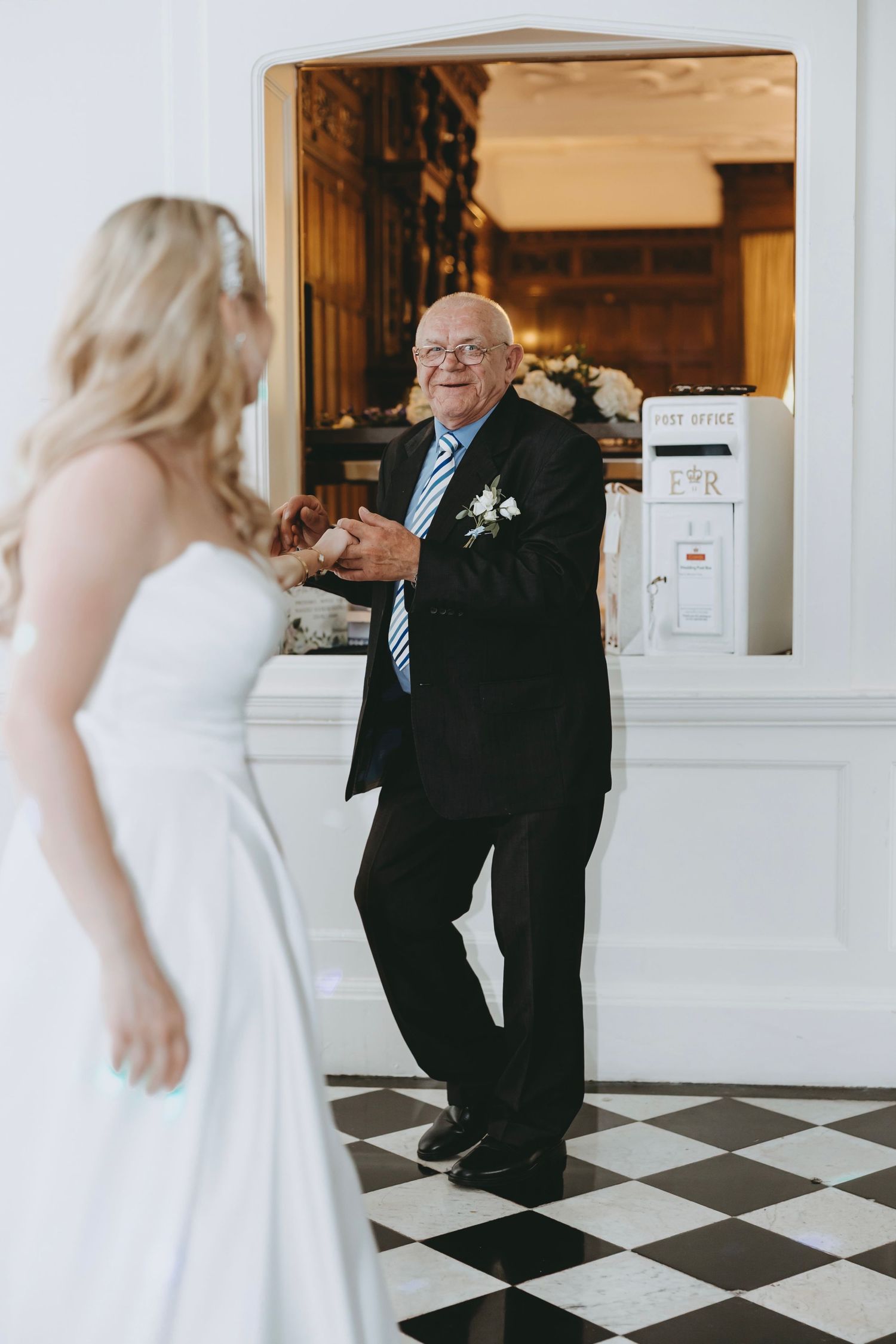 A touching first look moment between a bride in white and an elderly man in a black suit on a checkered floor.