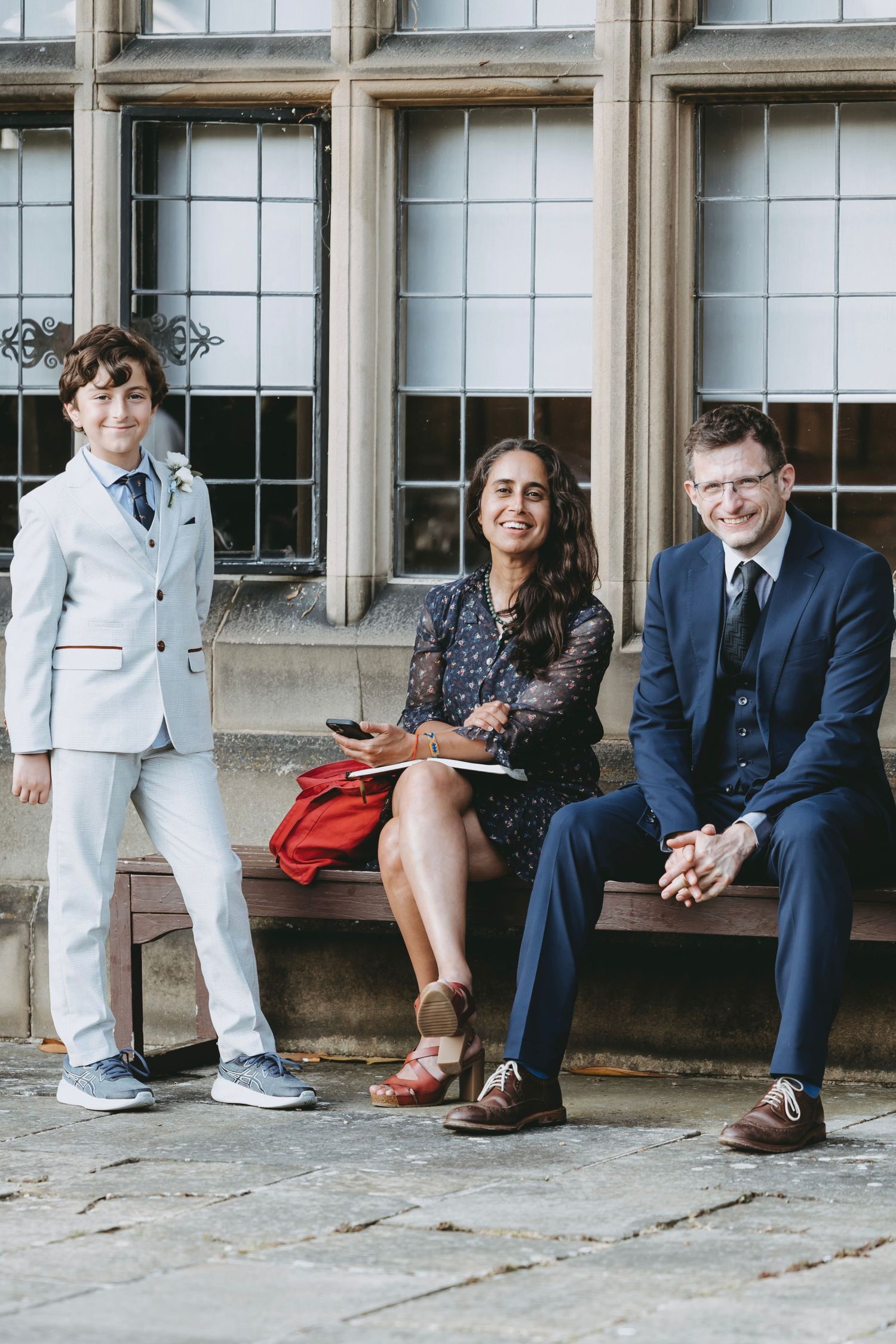Wedding party members sit casually on a stone ledge in front of large windows.