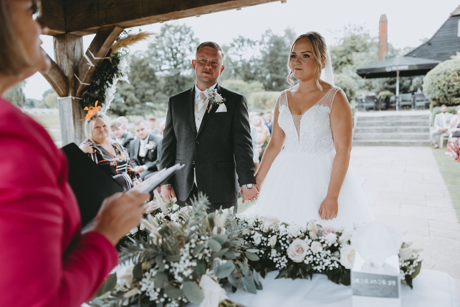 Wedding couple stands at decorated altar while officiant in pink conducts ceremony.