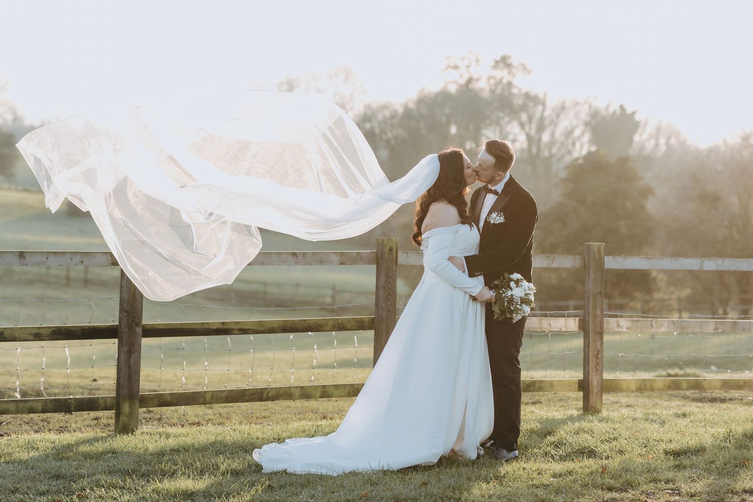 Romantic wedding couple embraces in a sunlit meadow as the bride's veil flows gracefully in the breeze.