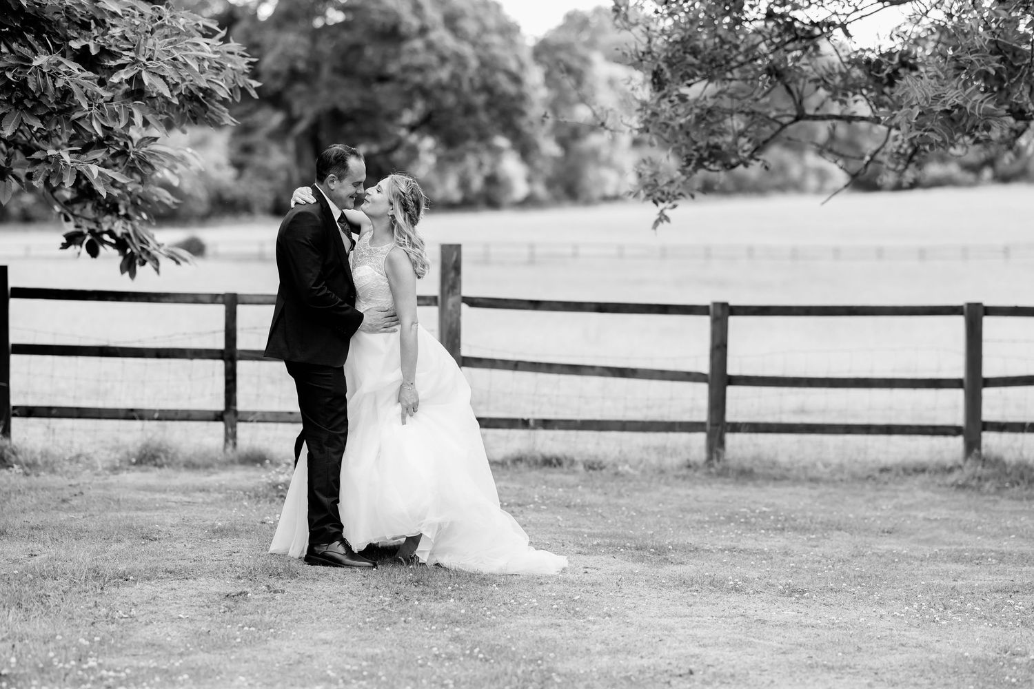 Black and white photo of a wedding couple by a white fence in an open field.