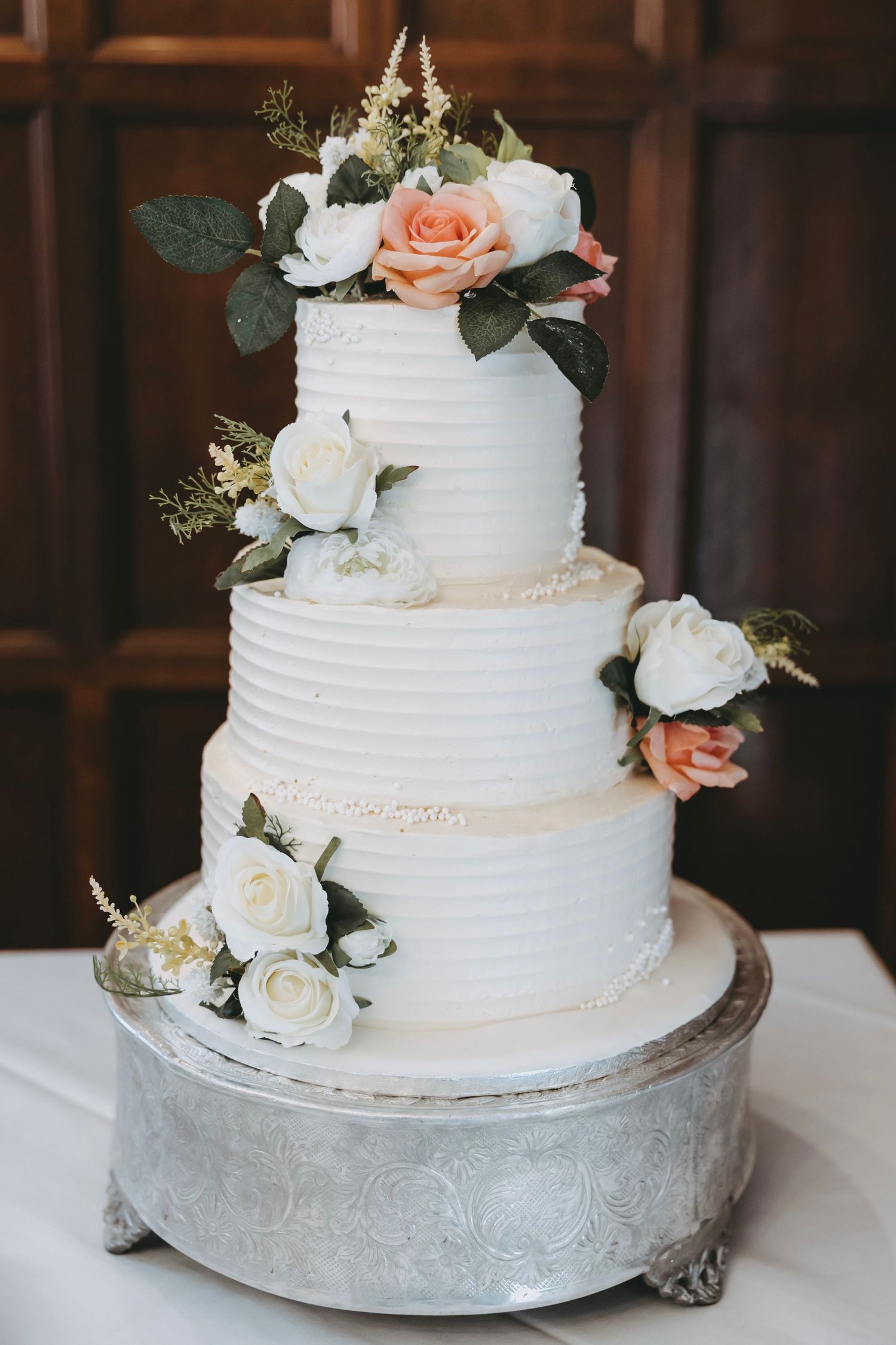 Elegant white wedding cake with peach and white roses displayed on silver cake stand.