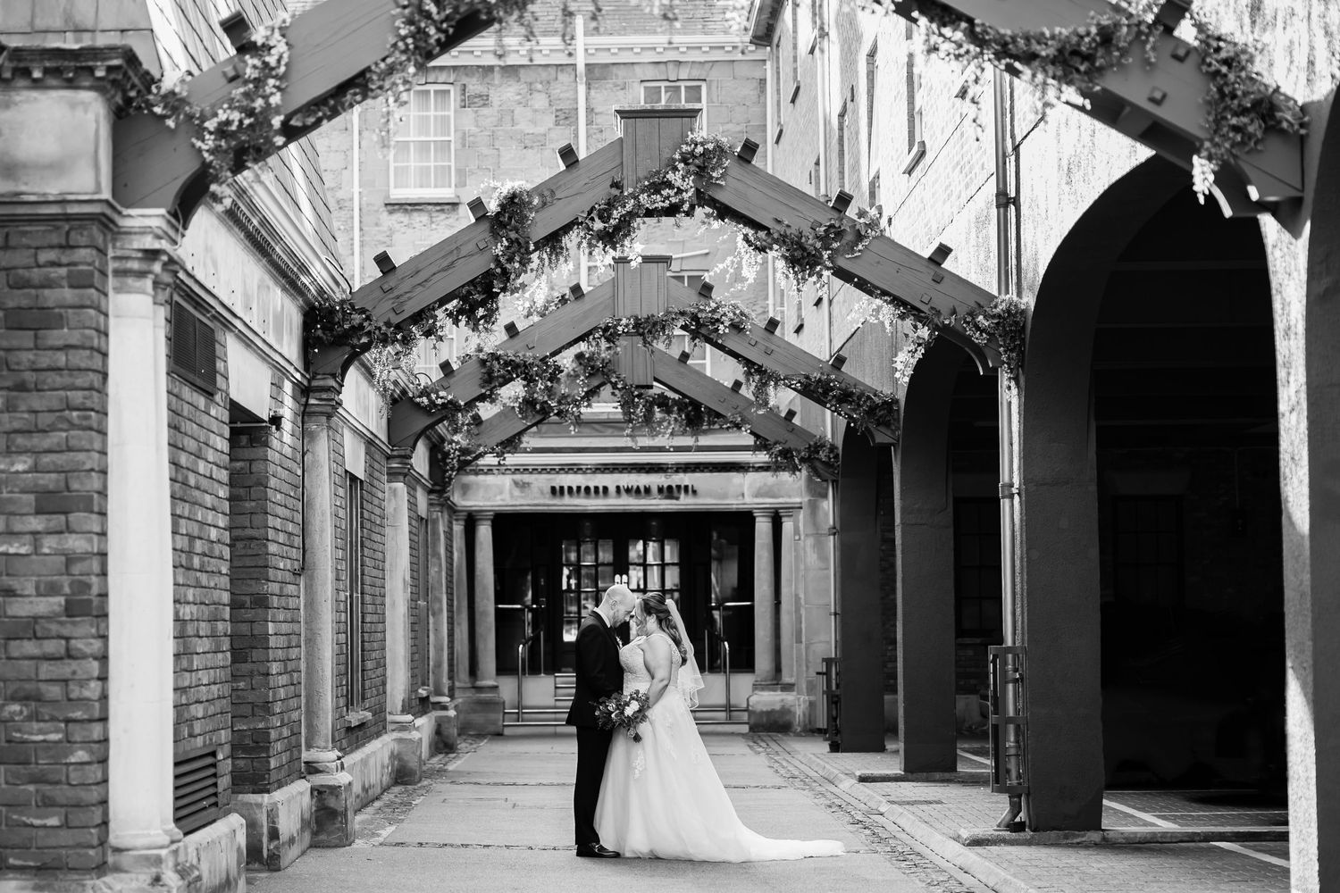 A black and white wedding photo of newlyweds under an archway with decorative lighting at The Swan Hotel, Bedford.