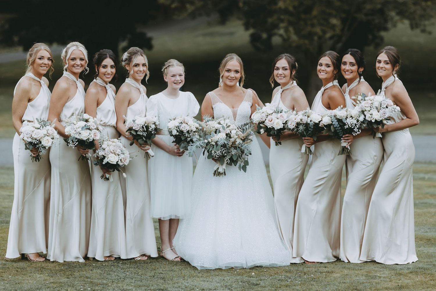 Bridesmaids in cream colored dresses pose together holding bouquets on grassy lawn.