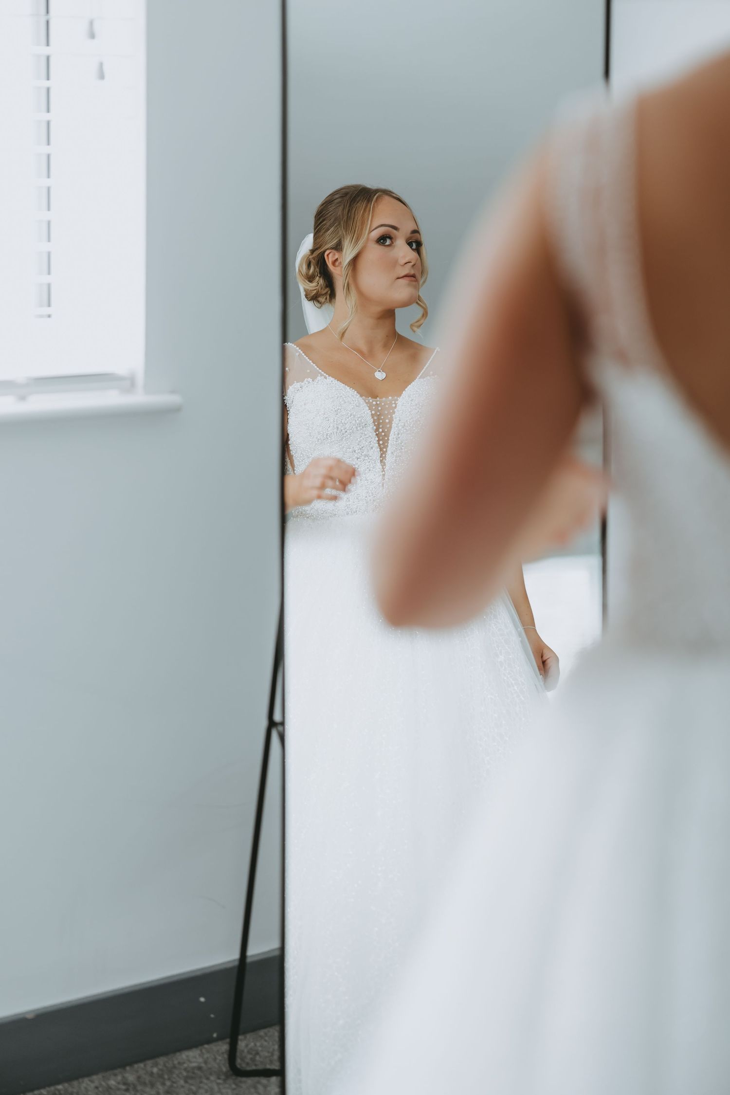 A bride in a white lace wedding gown looks at her reflection in a mirror during wedding preparations.