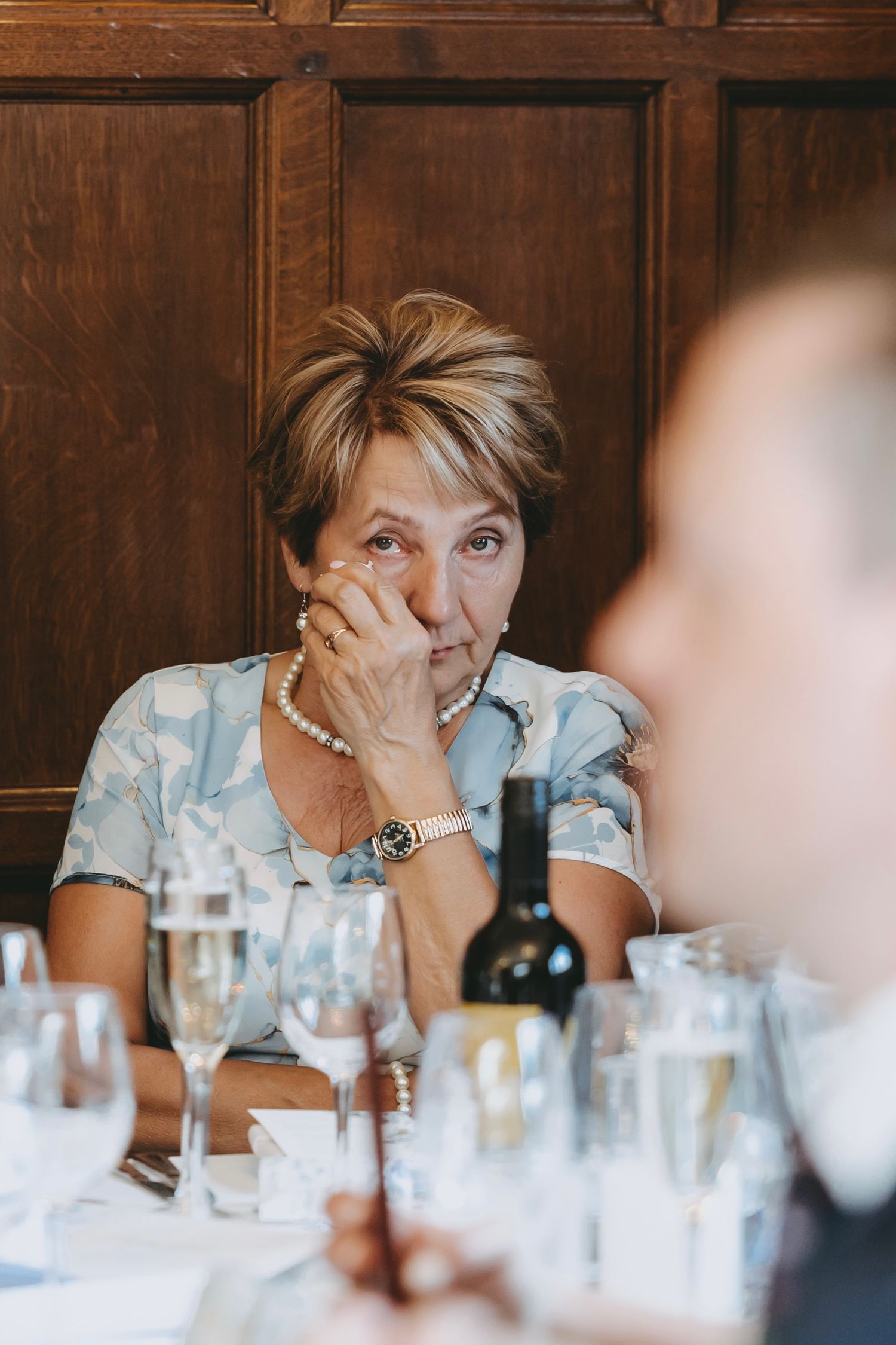 Guest at wedding reception seated at dining table with wine bottles and glassware.