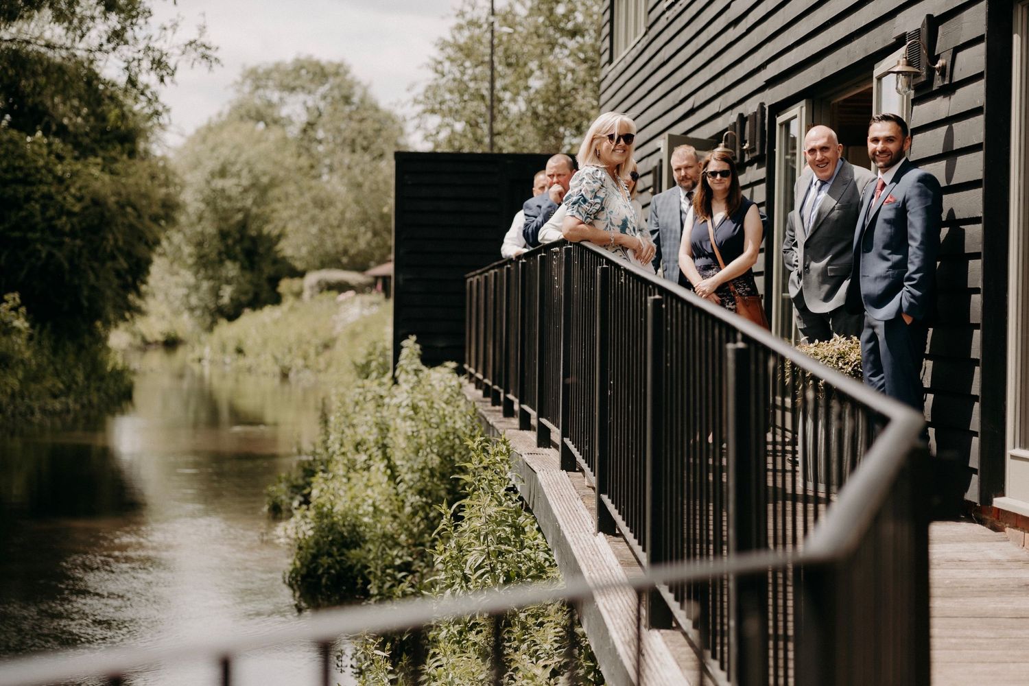 Wedding party members pose together on a wooden deck overlooking a serene pond.